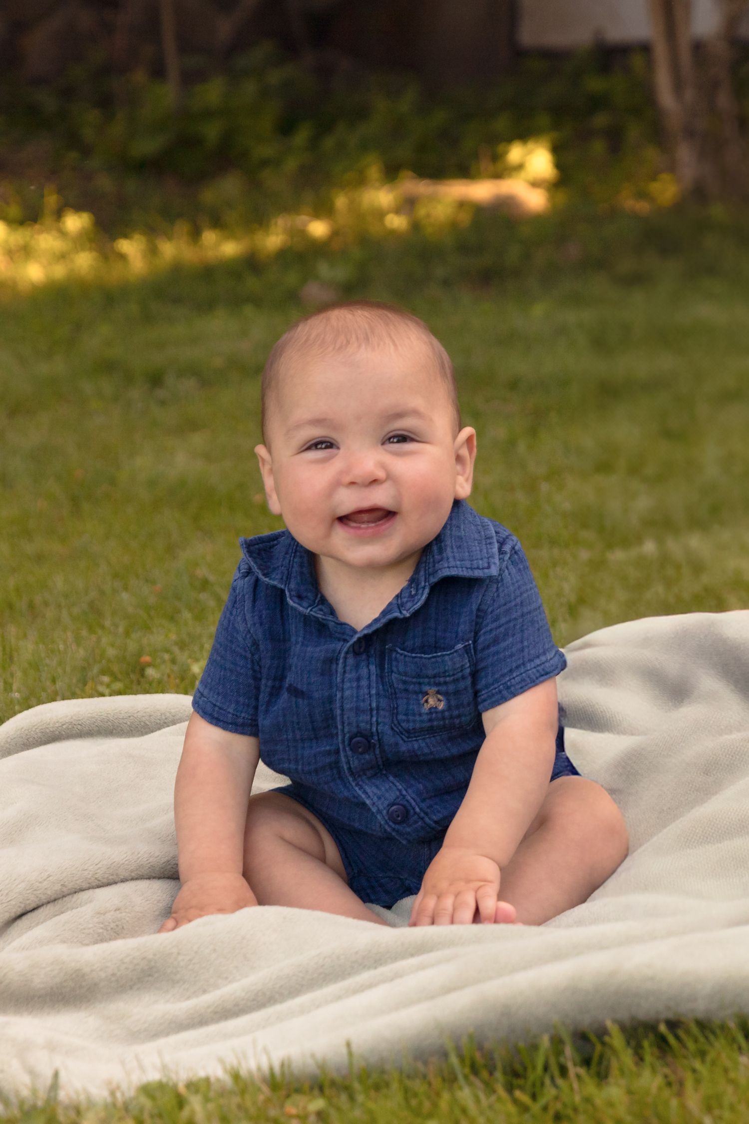 Baby in denim outfit smiling while lying on white blanket outdoors.