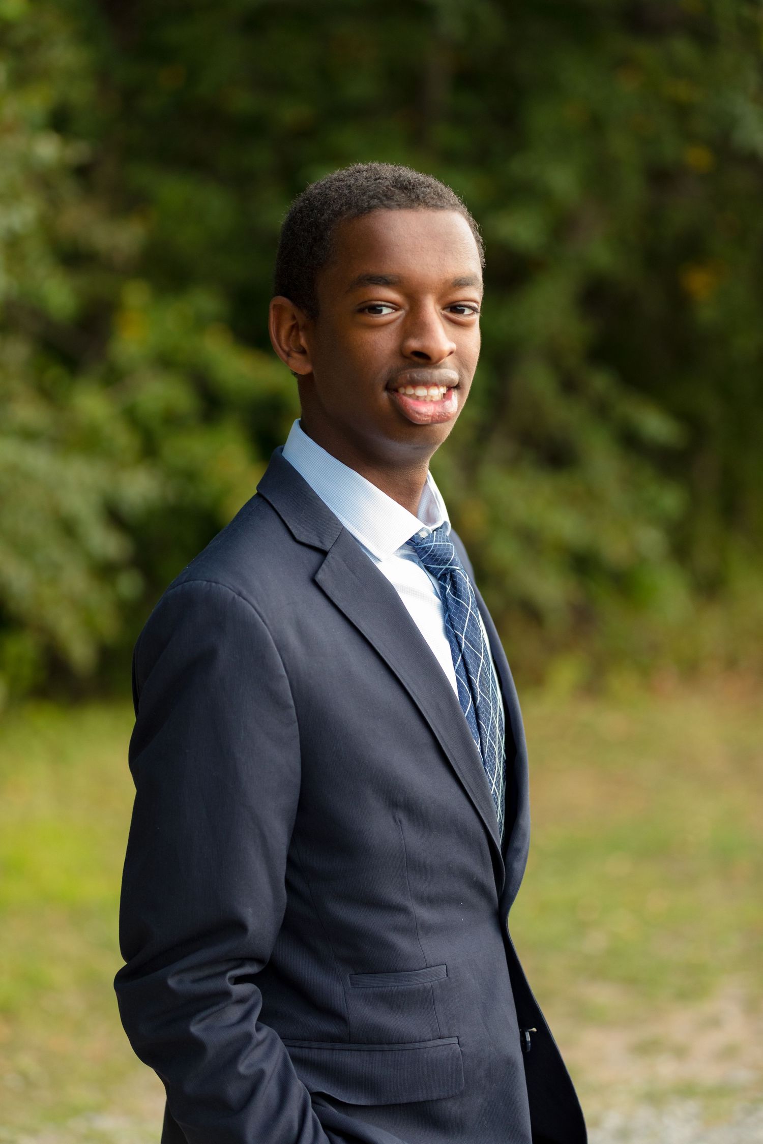 A person in formal business attire smiles while standing against a natural backdrop.