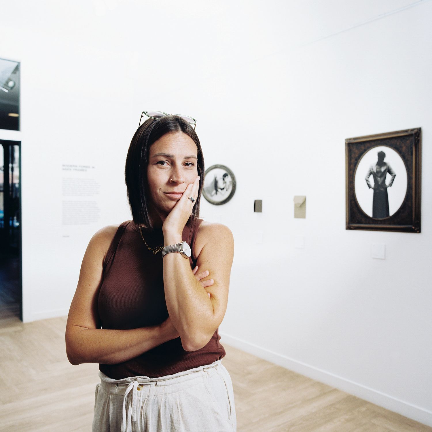 Person in a dark sleeveless top poses expressively in a minimalist gallery space with framed artwork on white walls.