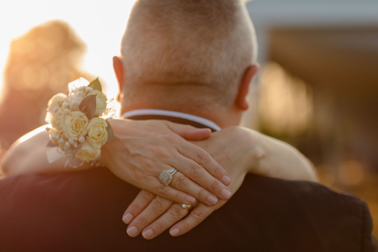 Close-up of hands with wedding rings and a white floral wrist corsage in golden sunlight.
