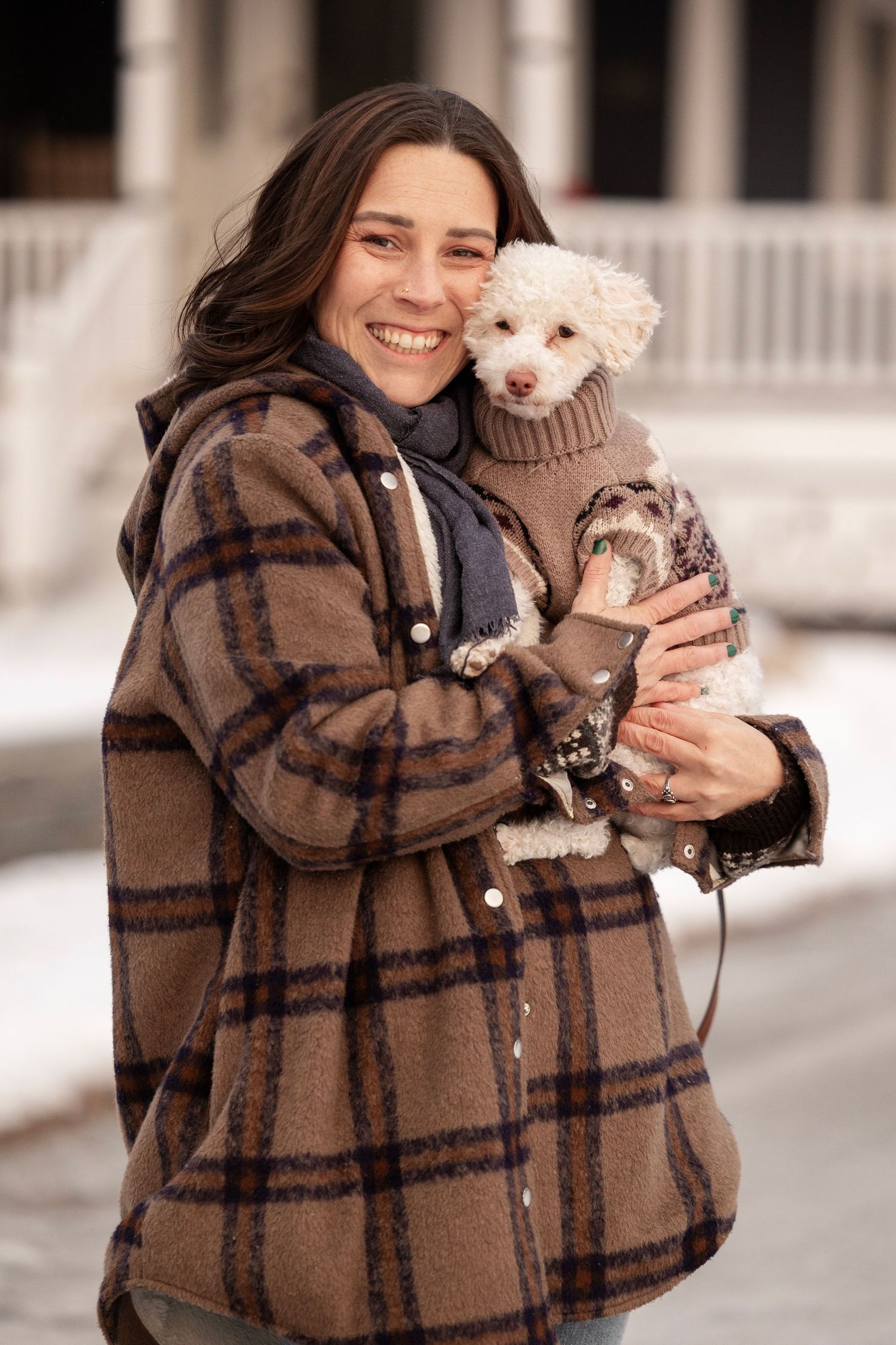 Person cuddling small white dog while wearing cozy plaid jacket outdoors.