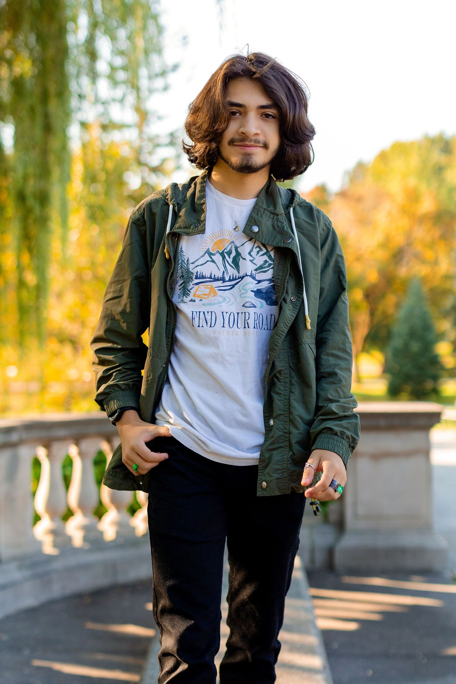 Person wearing green jacket and white t-shirt poses outdoors against autumn trees and willow branches in sunlight.