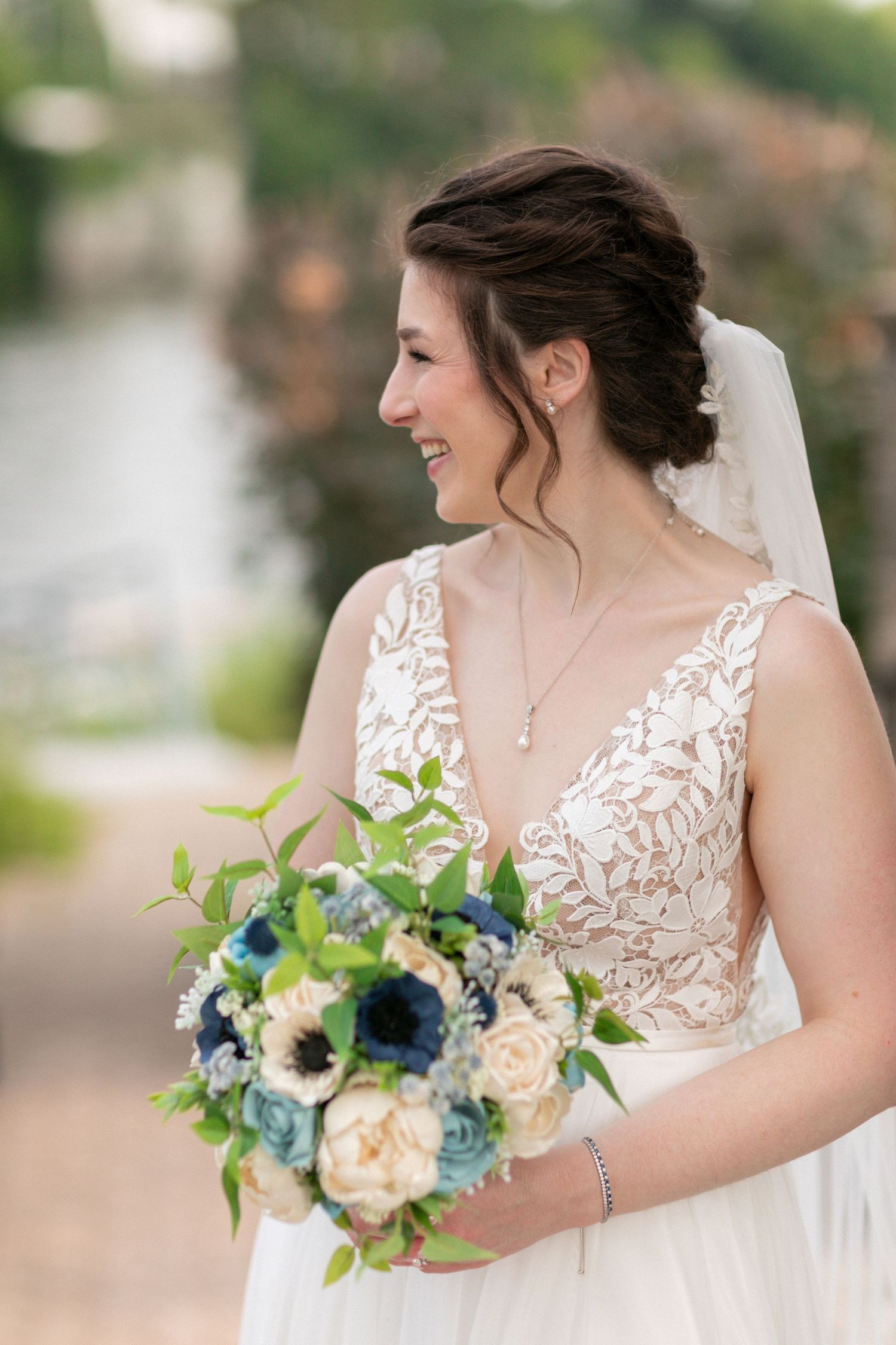 A bride in a lace detailed dress holds a blue and white bouquet while smiling.