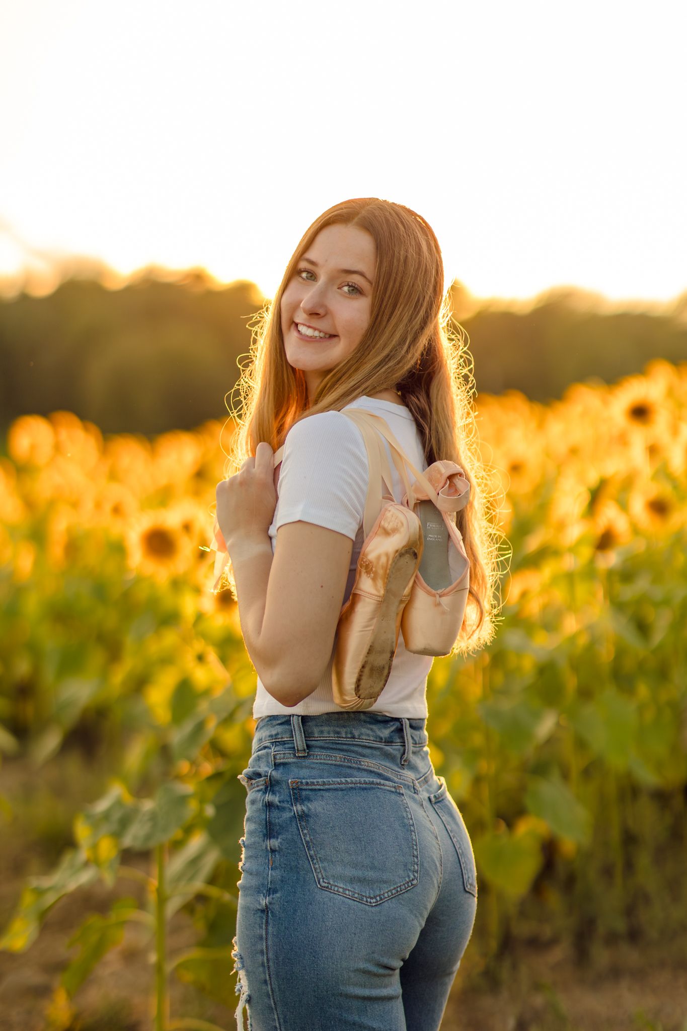 A Ballerina in jeans and white shirt poses playfully in a sunlit vineyard at golden hour holding her pointe shoes.