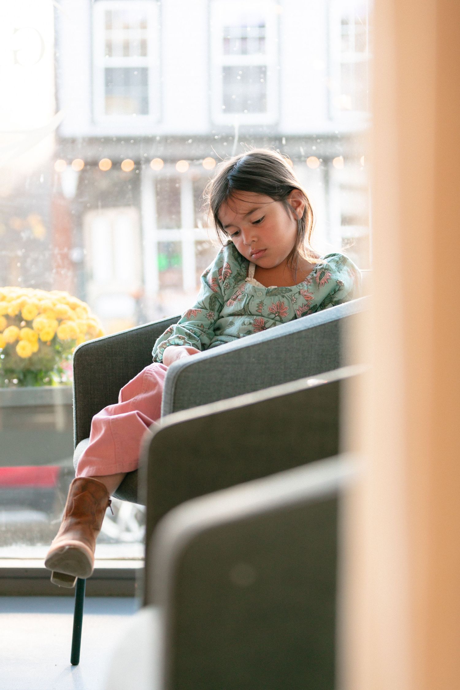 Young person resting on a grey couch near a window with yellow flowers visible outside.