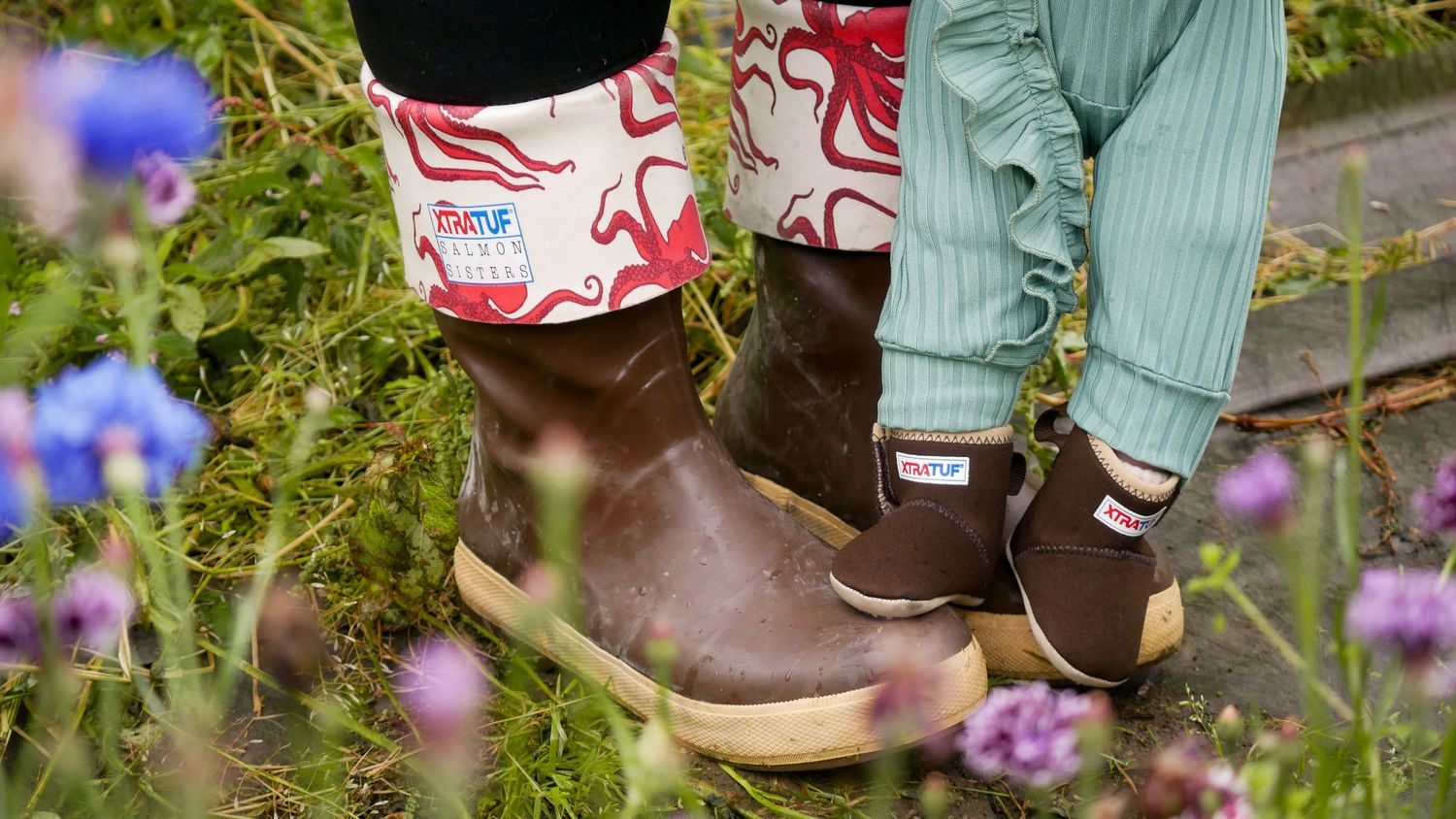 A person wearing Xtratuf boots walks along a garden path with blooming purple flowers with baby wearing matching boots.
