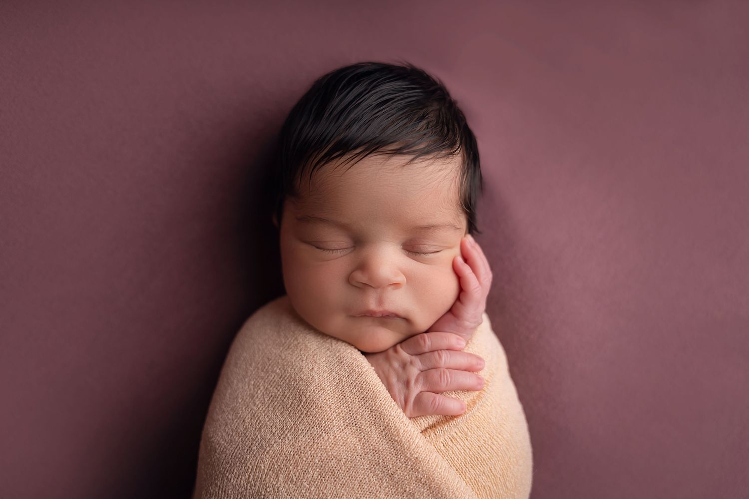 Close-up image of a newborn girl swaddled in a peach colored wrap against a plum colored backdrop during a Chapin SC Studio Newborn Session.