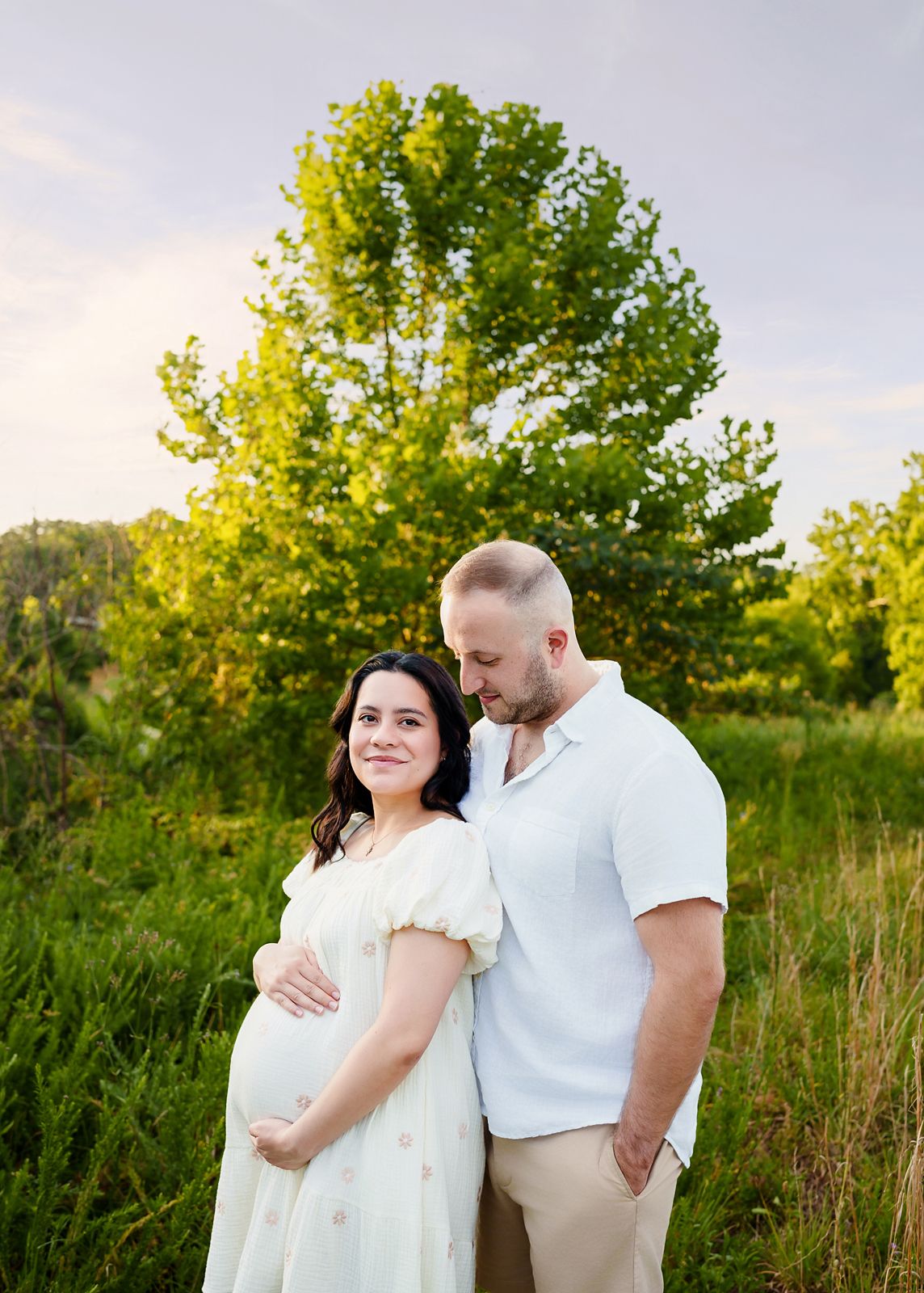 Mom and Dad in light color clothing against a lush green field with sunlight hitting a tree behind them in a Columbia SC Maternity Session.