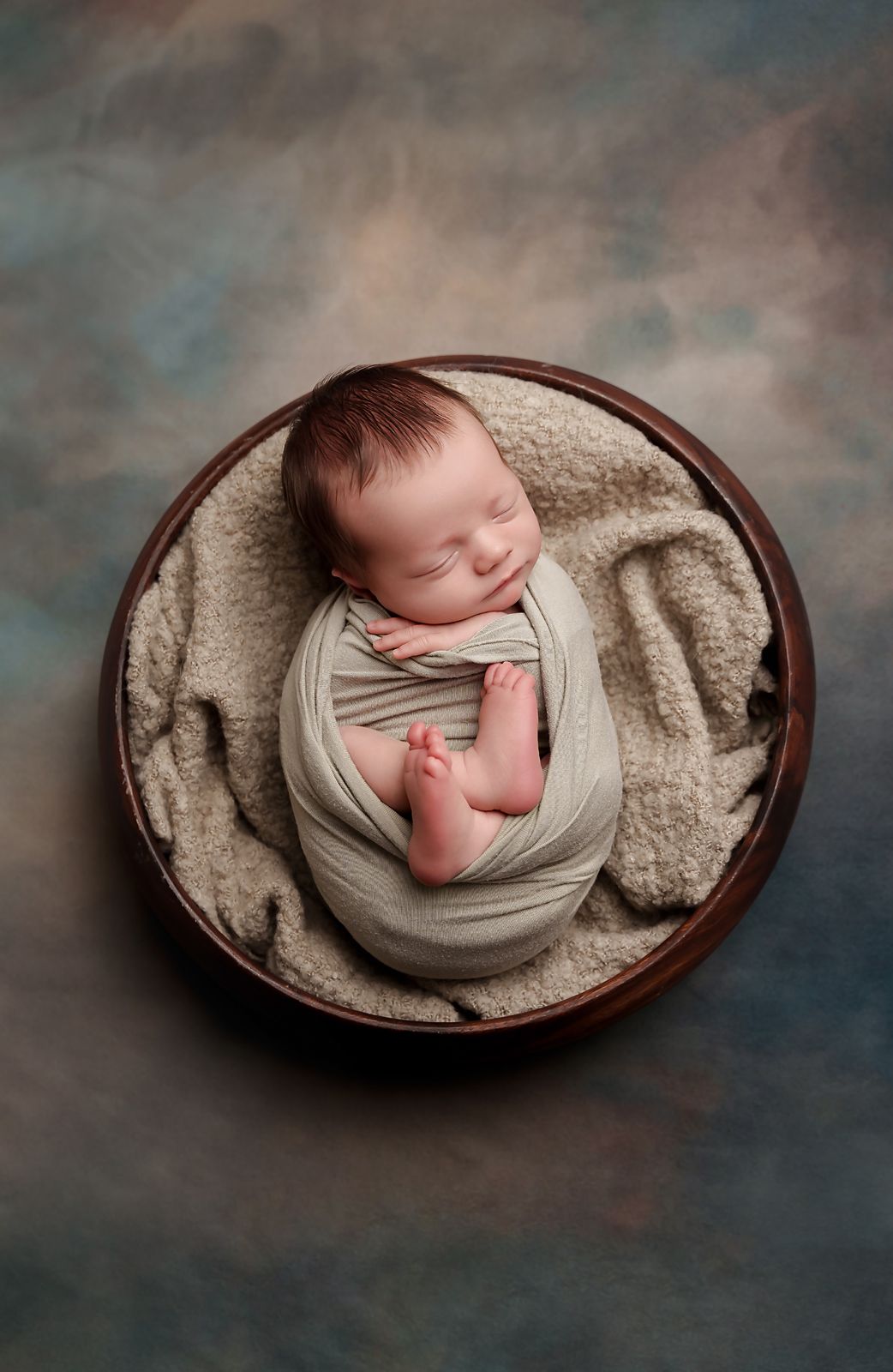 Newborn boy swaddled in a sage green wrap and posed in a round prop against a darker green and tan backdrop during a Columbia SC Session.