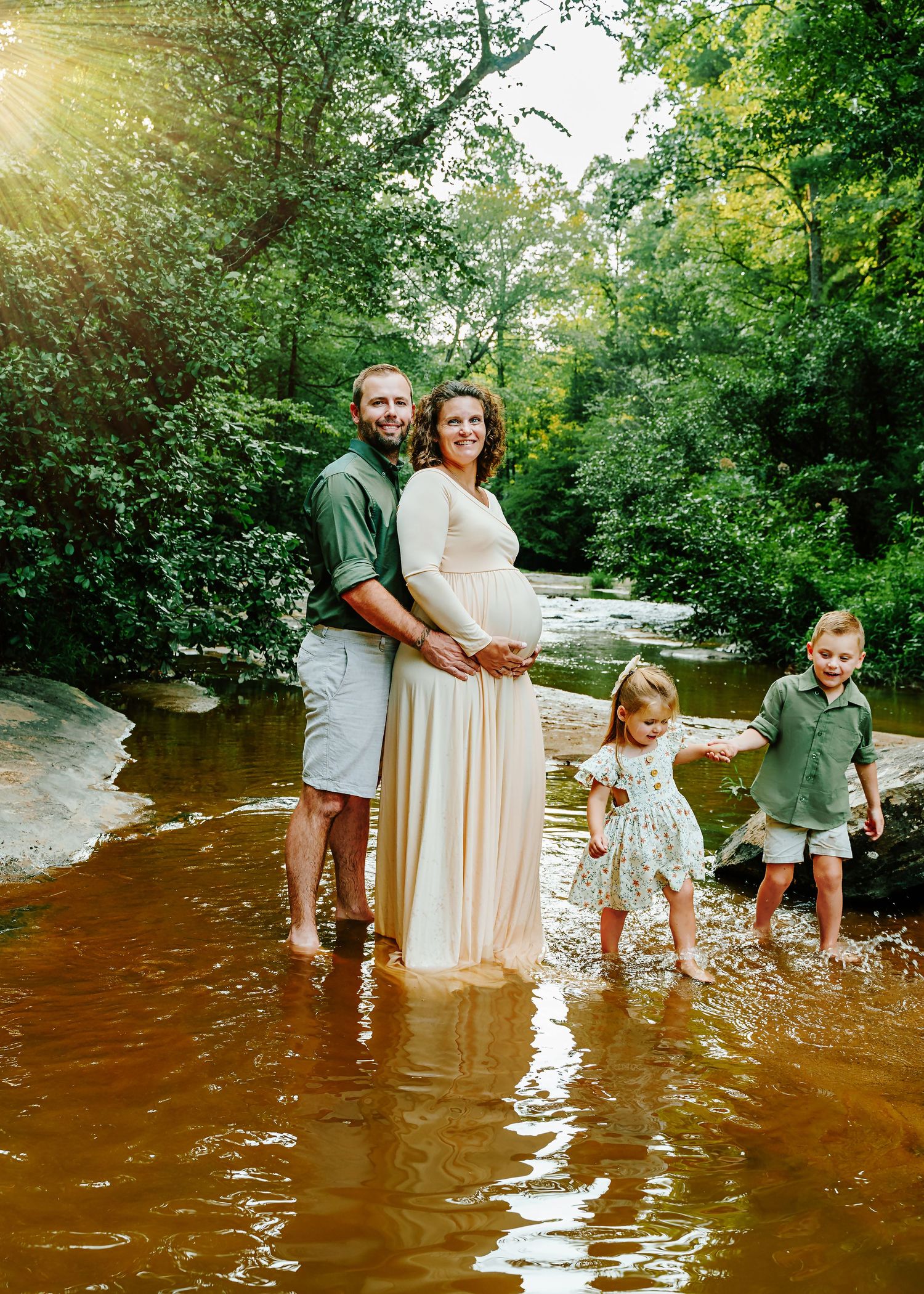 Dad embracing Mom as their kids hold hands and run around them during an Upstate SC creek Maternity Portrait Session.
