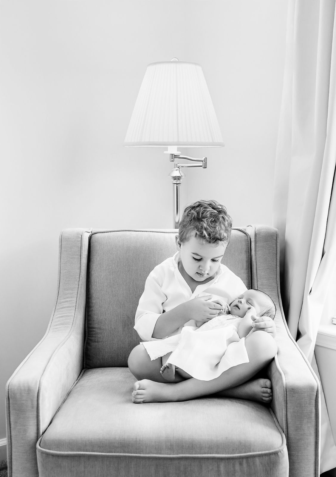 Sweet black and white image of a young boy sitting in a chair and holding his baby sister during an Irmo SC Lifestyle Newborn Session.