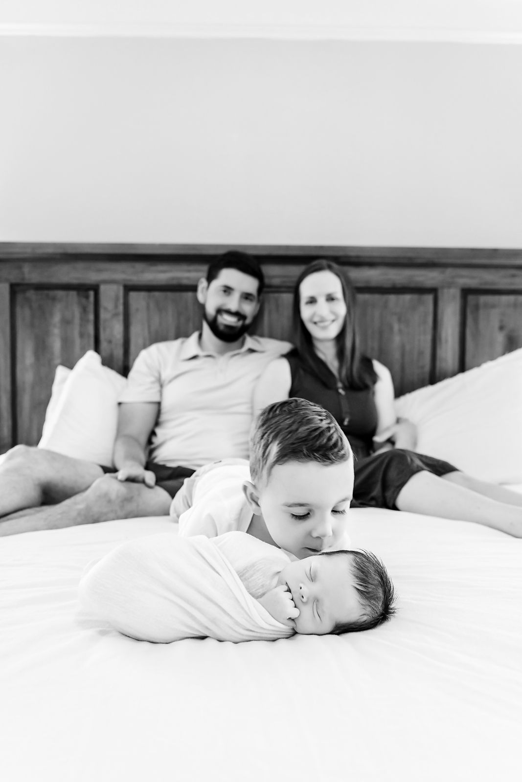 A toddler kisses his newborn sibling on the forehead as Mom and Dad look from the background during a Lexington SC in-home newborn session.