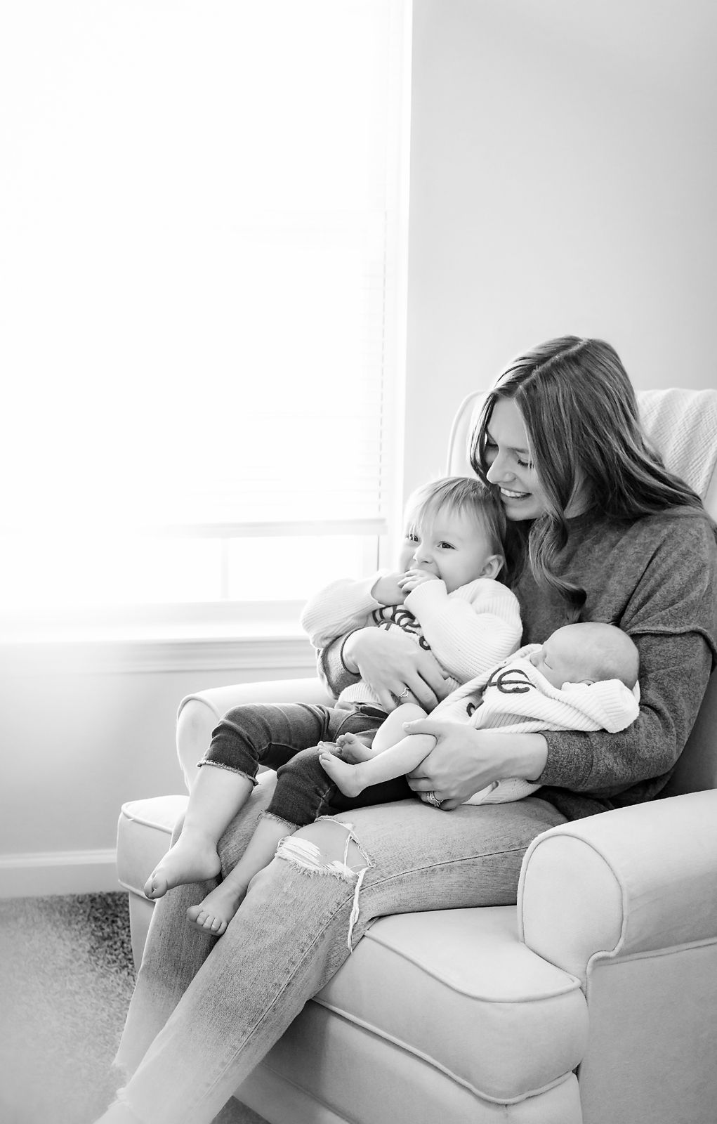 Mom sitting in a chair by the window with both of her babies and lovingly looking down at them during a Columbia SC In-Home Newborn Session.