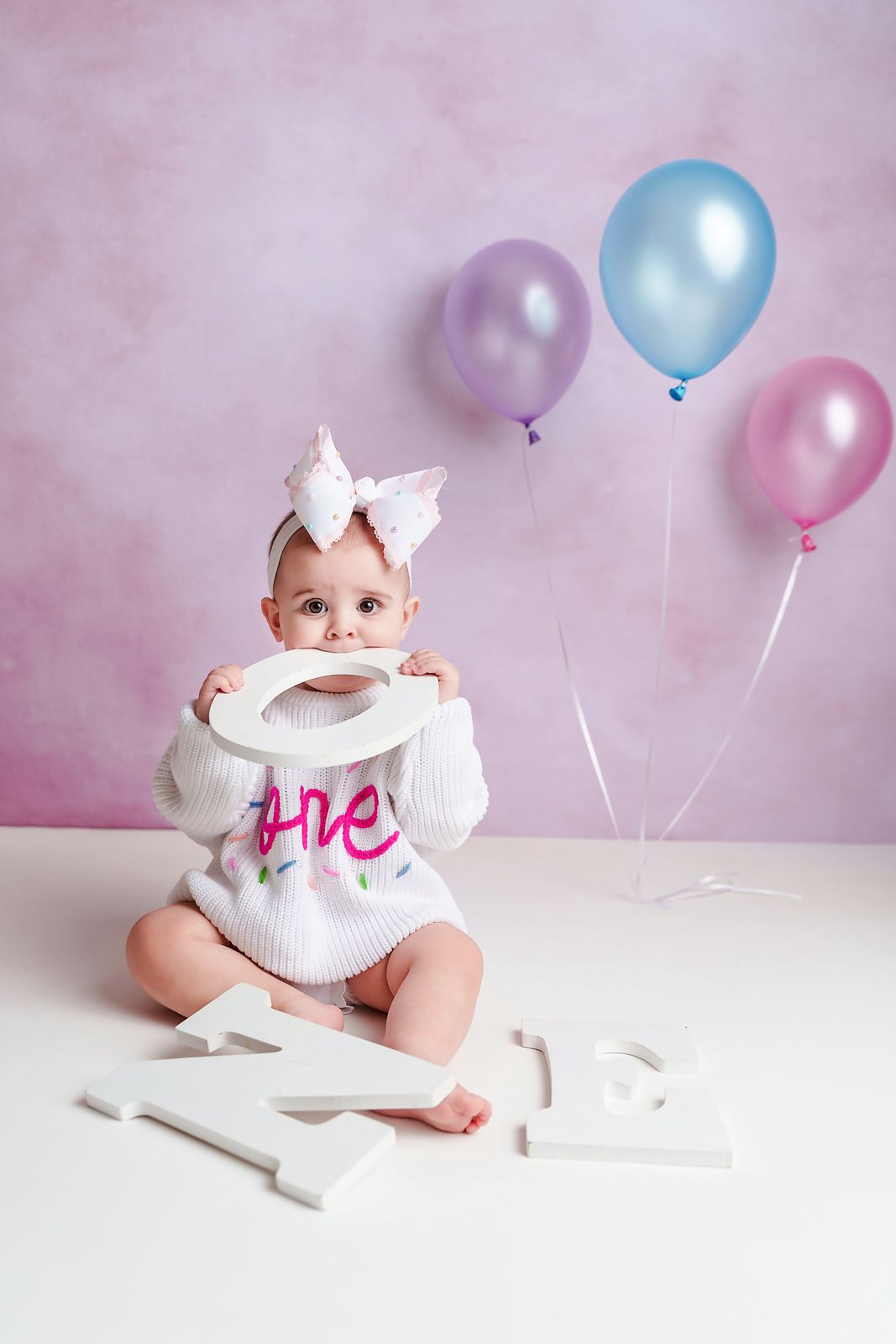 Simple Newberry SC 1st Birthday cake smash session of a girl in a white ONE sweater against a pink backdrop with a few pastel balloons.