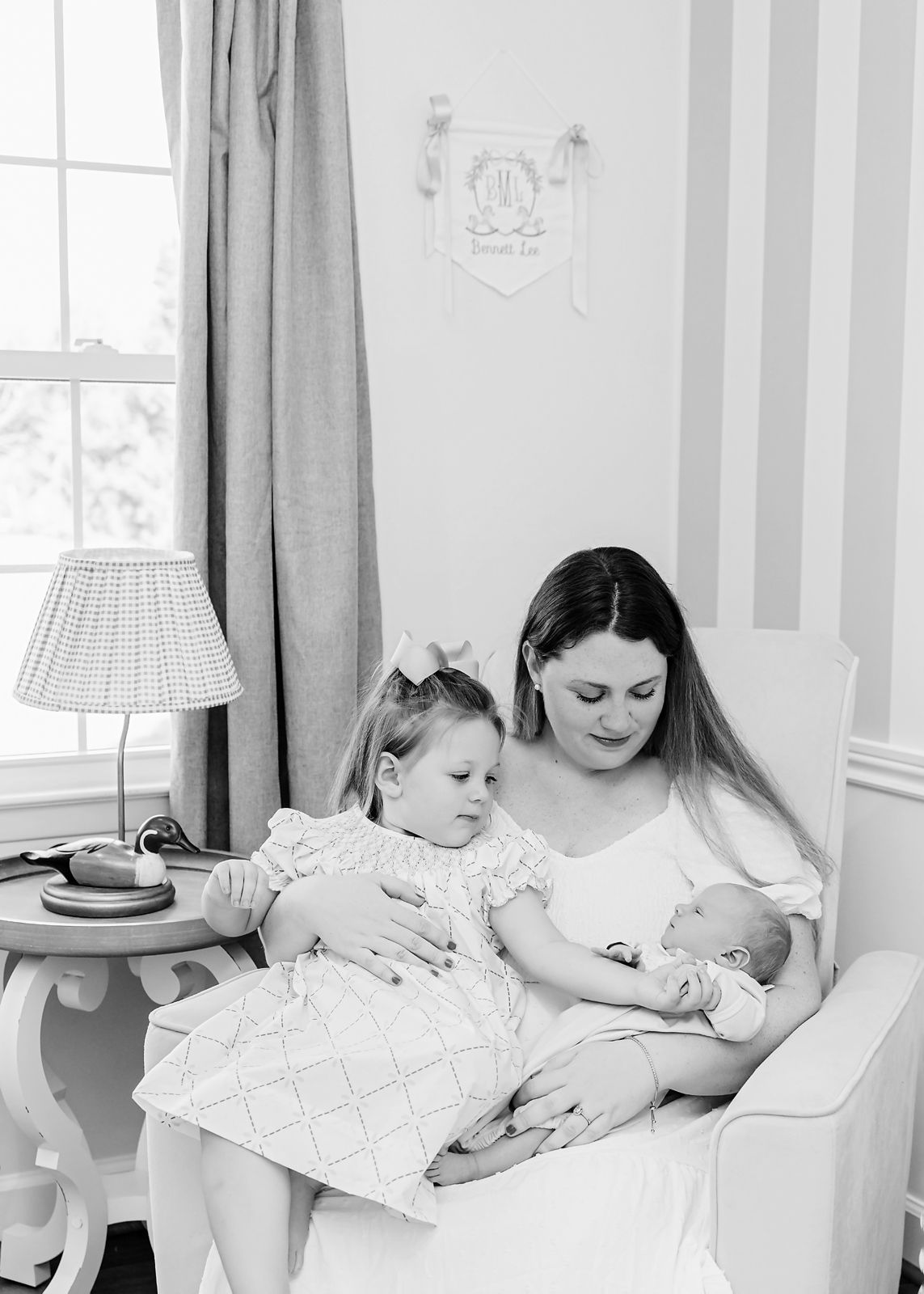 Newberry SC in home newborn session: mom sitting in baby nursery cuddling both her newborn and toddler as the toddler holds babies hand.