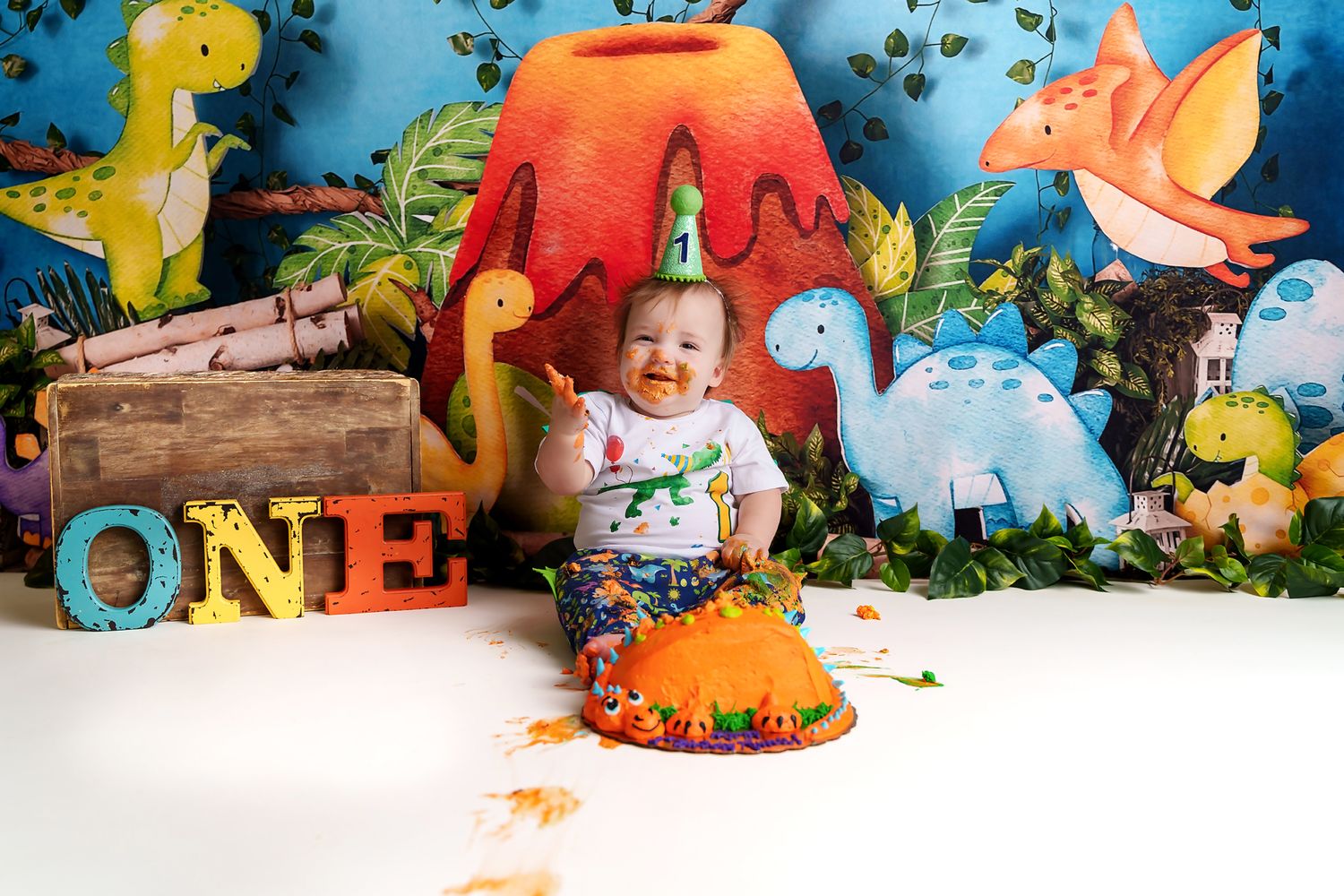One year old little boy smiling while destroying his cake in front of a colorful dinosaurs themed backdrop during a Chapin SC Cake Smash.