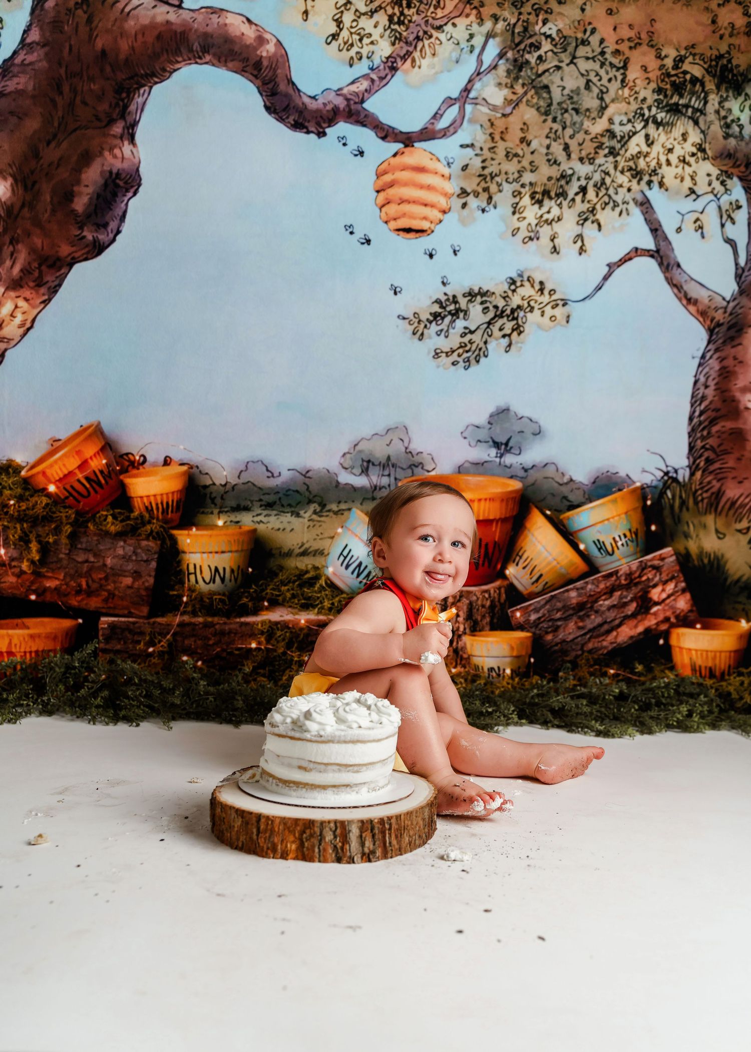 Adorable little one year old boy grinning at the camera during his Pooh Hunny Pot themed 1st Birthday Cake Smash Session in Chapin SC.