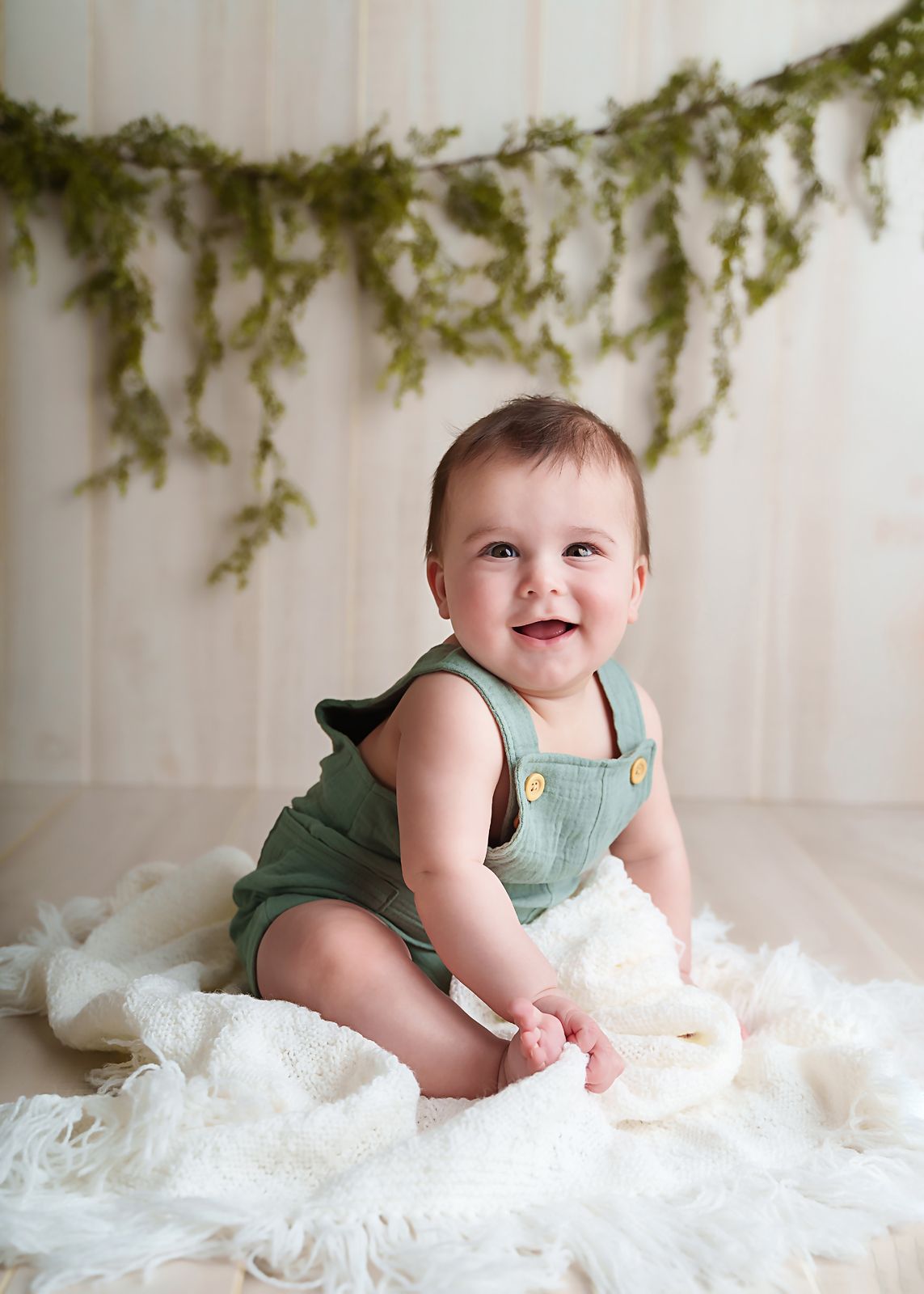 Lexington SC studio portrait of a 6 month old baby boy sitting on a white blanket with greenery hanging in the background.