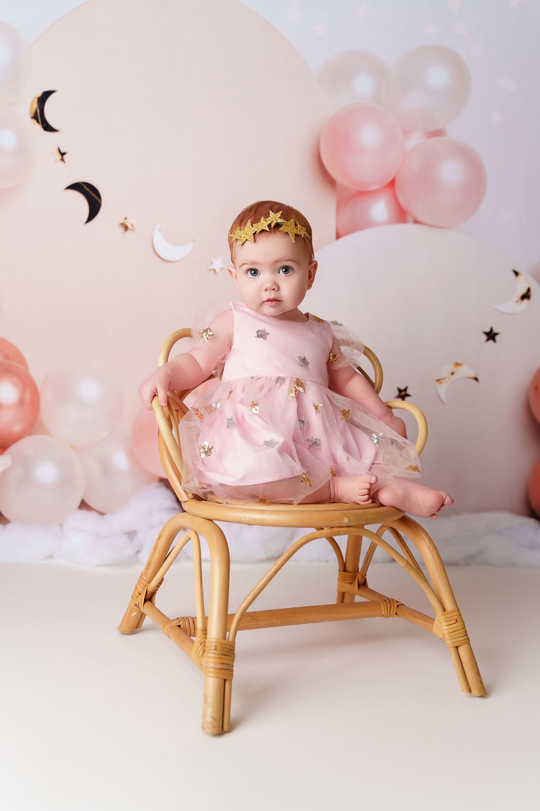 Adorable 1 yr old girl sitting in a small chair in front of a pink and gold celestial themed backdrop during her Lexington SC Cake Smash.