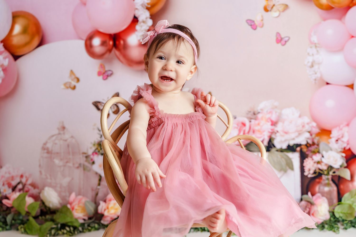 Little girl in a pink dress smiling at the camera in front of a floral and butterfly themed backdrop during her 1st Birthday Cake Smash.