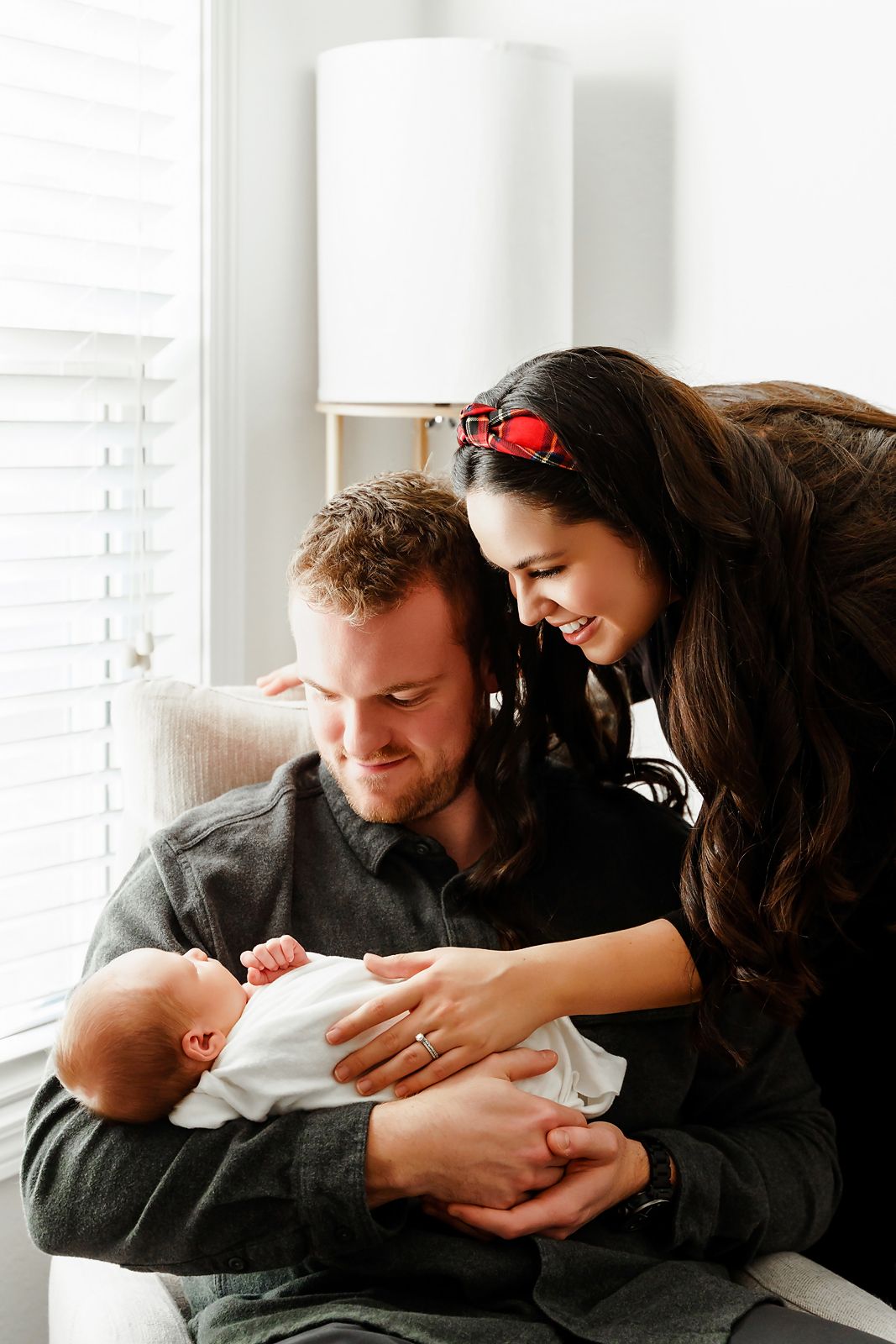 Dad holding his newborn baby in a chair as mom loving leans down to embrace them during a Lexington SC in-home newborn portrait session.