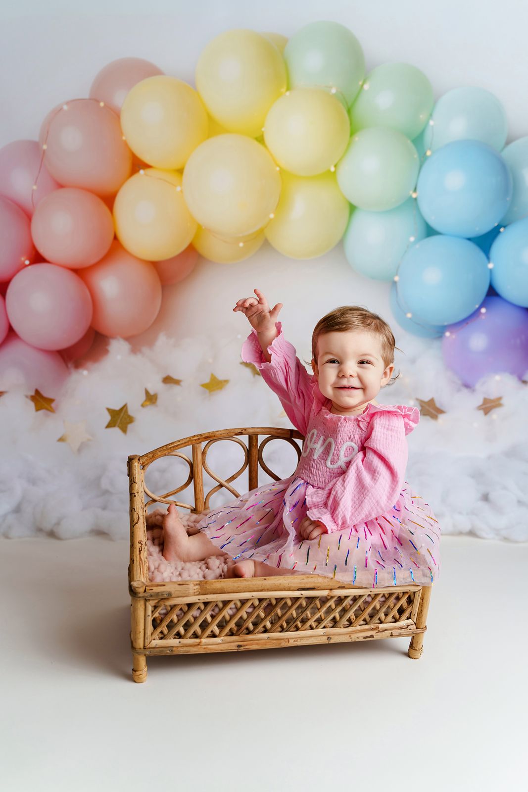 Adorable 1st Birthday Portrait of a girl sitting on a bamboo prop in front of a pastel rainbow balloon arch during a Chapin SC Cake Smash.