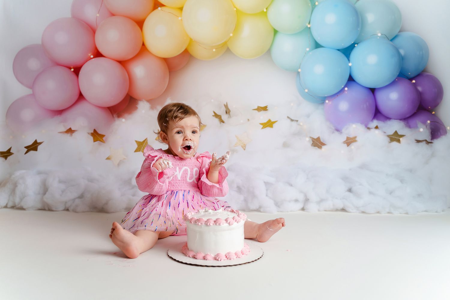 Adorable little girl enjoying her cake smash in front of a pastel rainbow balloon arch during her Chapin SC Cake Smash Portrait Session.