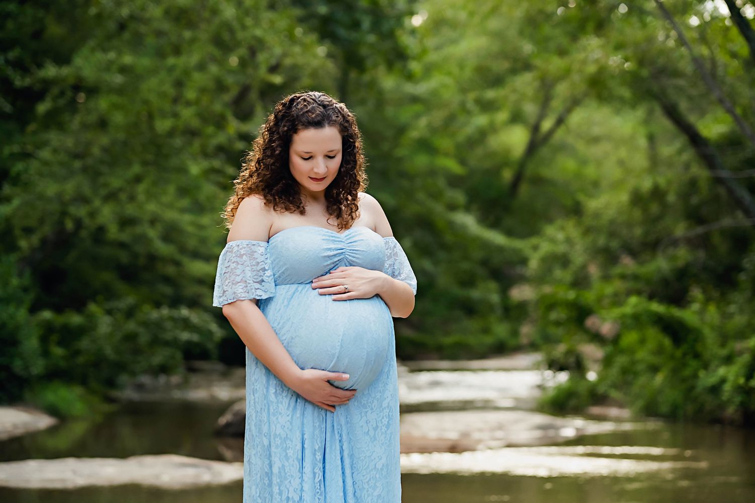 Mom to be in a blue lace dress cradling her belly in front of a stunning creek setting with greenery during an Upstate SC Maternity Session.