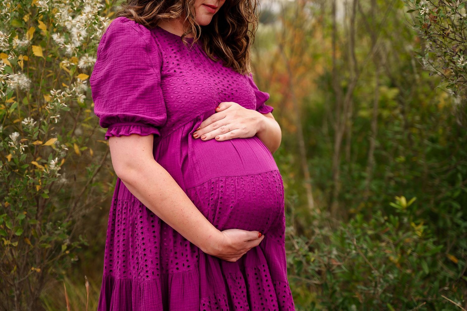 Close-up image of Mom to be cradling her belly in a purple dress standing in front of a background of muted greenery taken in Columbia SC.