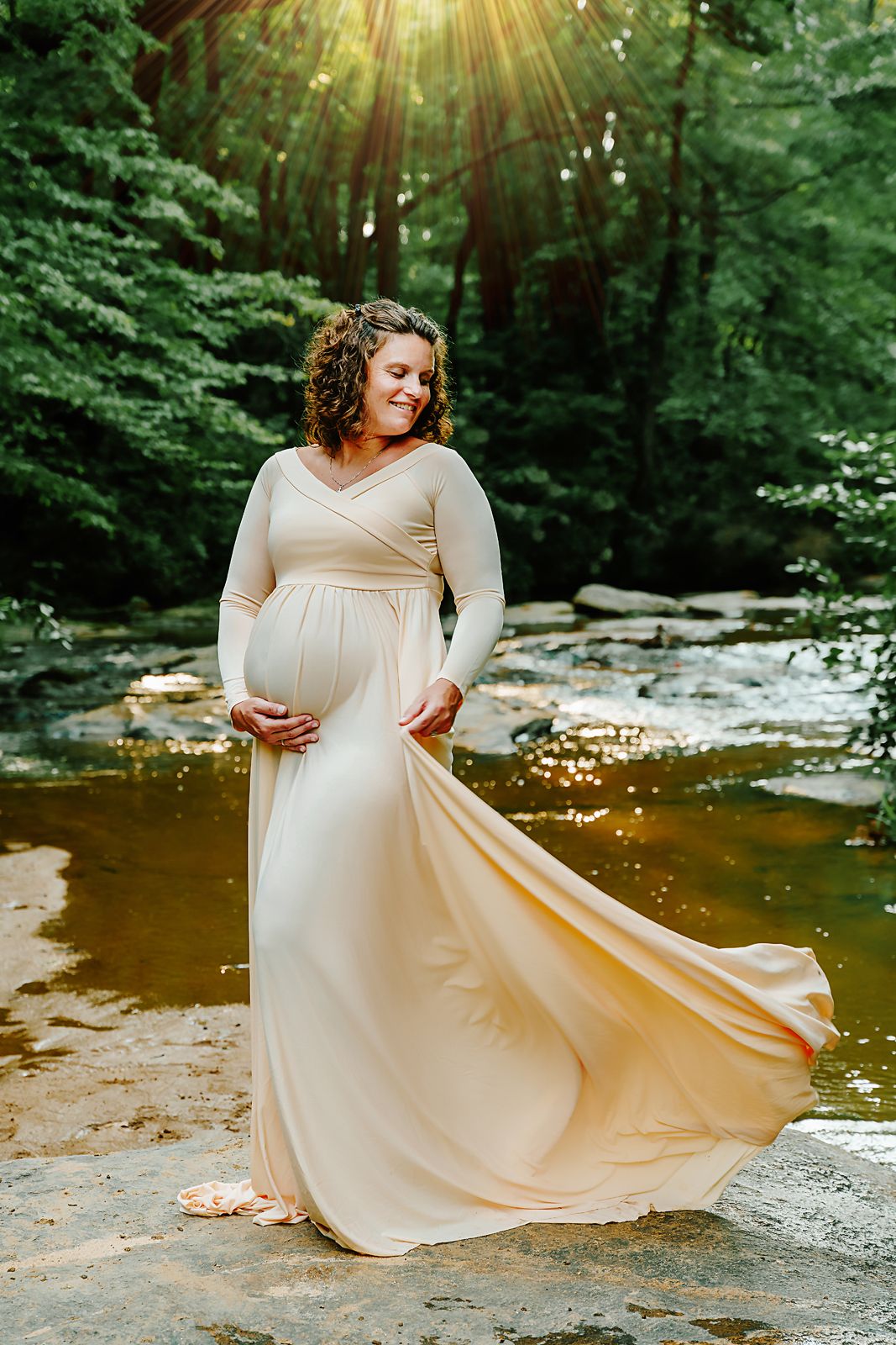 Mom to be in a cream colored dress playfully swirling the bottom during a Maternity Session at a creek in Upstate SC.