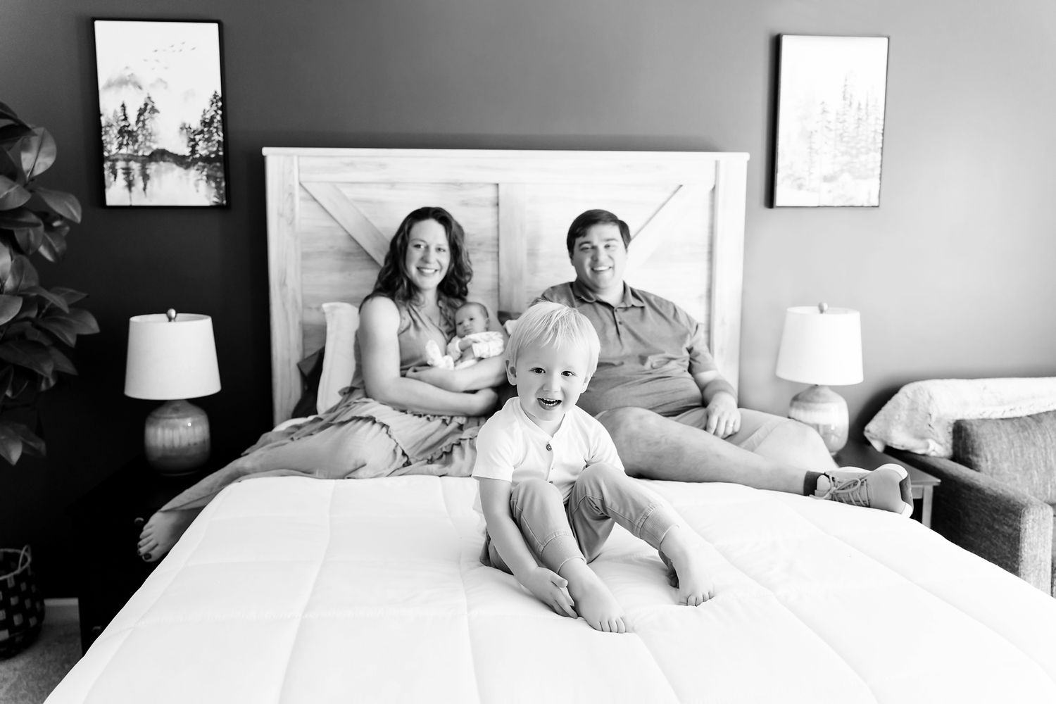 Toddler sitting at front of bed while mom and dad snuggle the newborn during an in-home newborn portrait session in Ballantine SC.