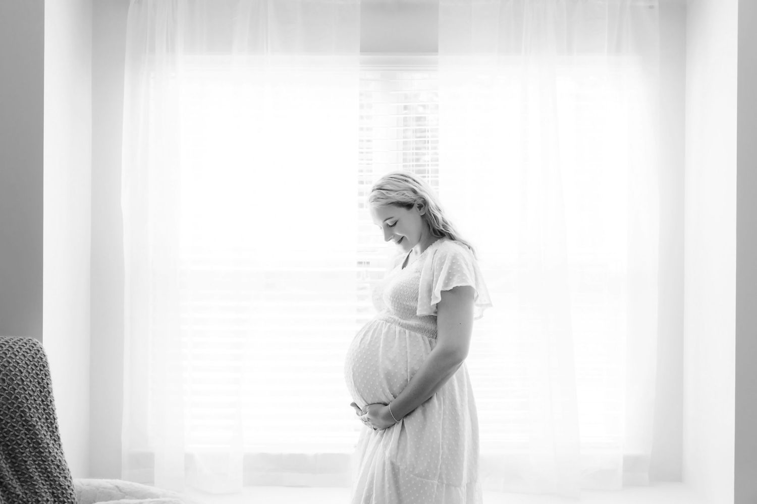 Black and white image of mom to be standing in front of a window while looking down at her belly in a Blythewood In-Home Maternity Session.