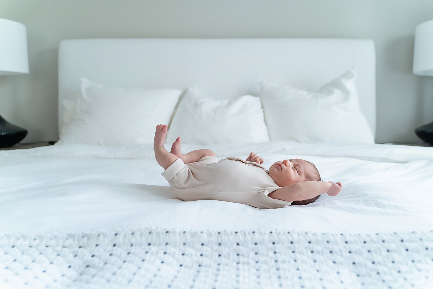 Newborn baby in a tan romper stretching on a bed made with a white duvet during a Chapin SC lifestyle in-home newborn portrait session.