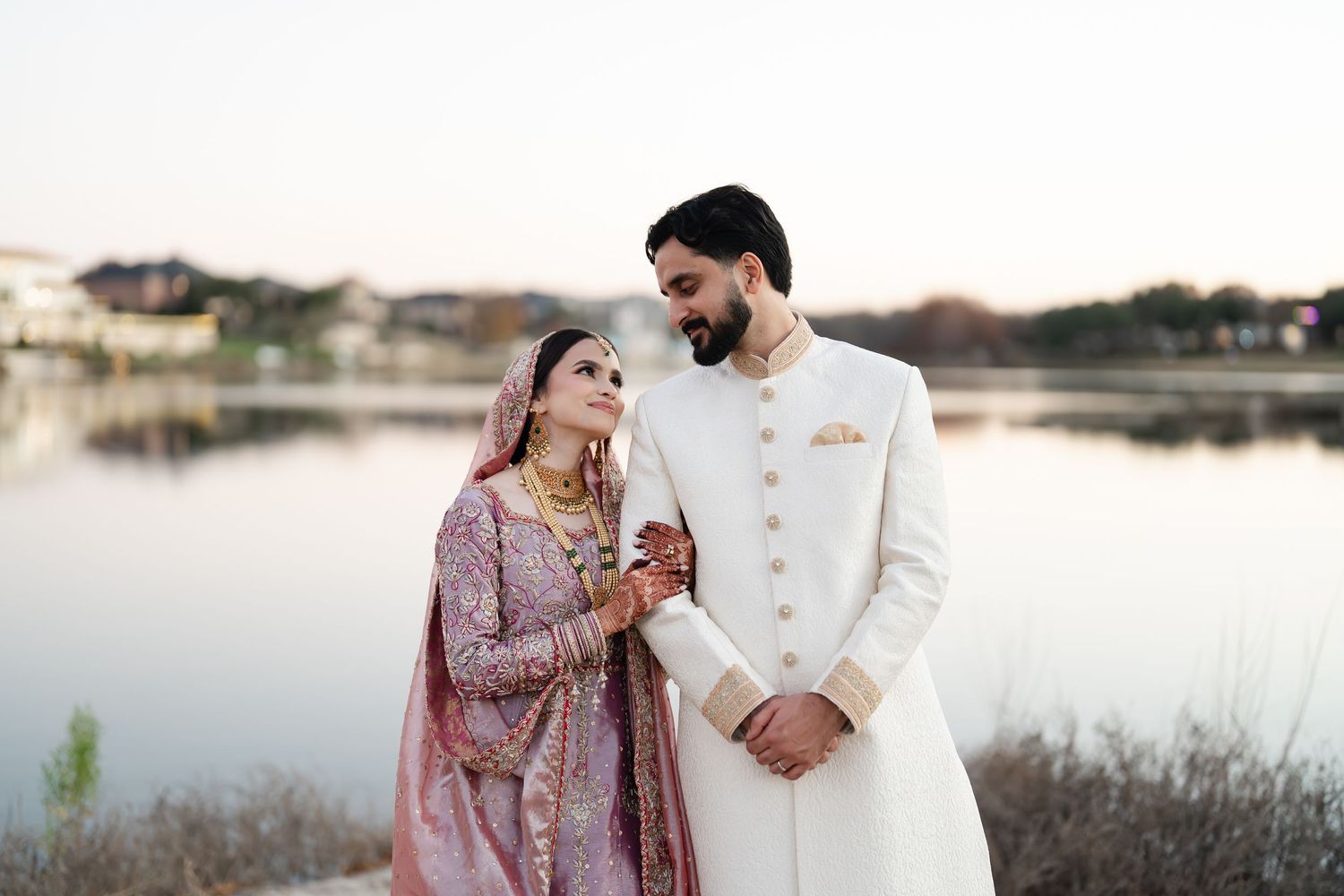 Couple in traditional South Asian wedding attire pose together at sunset by lake.