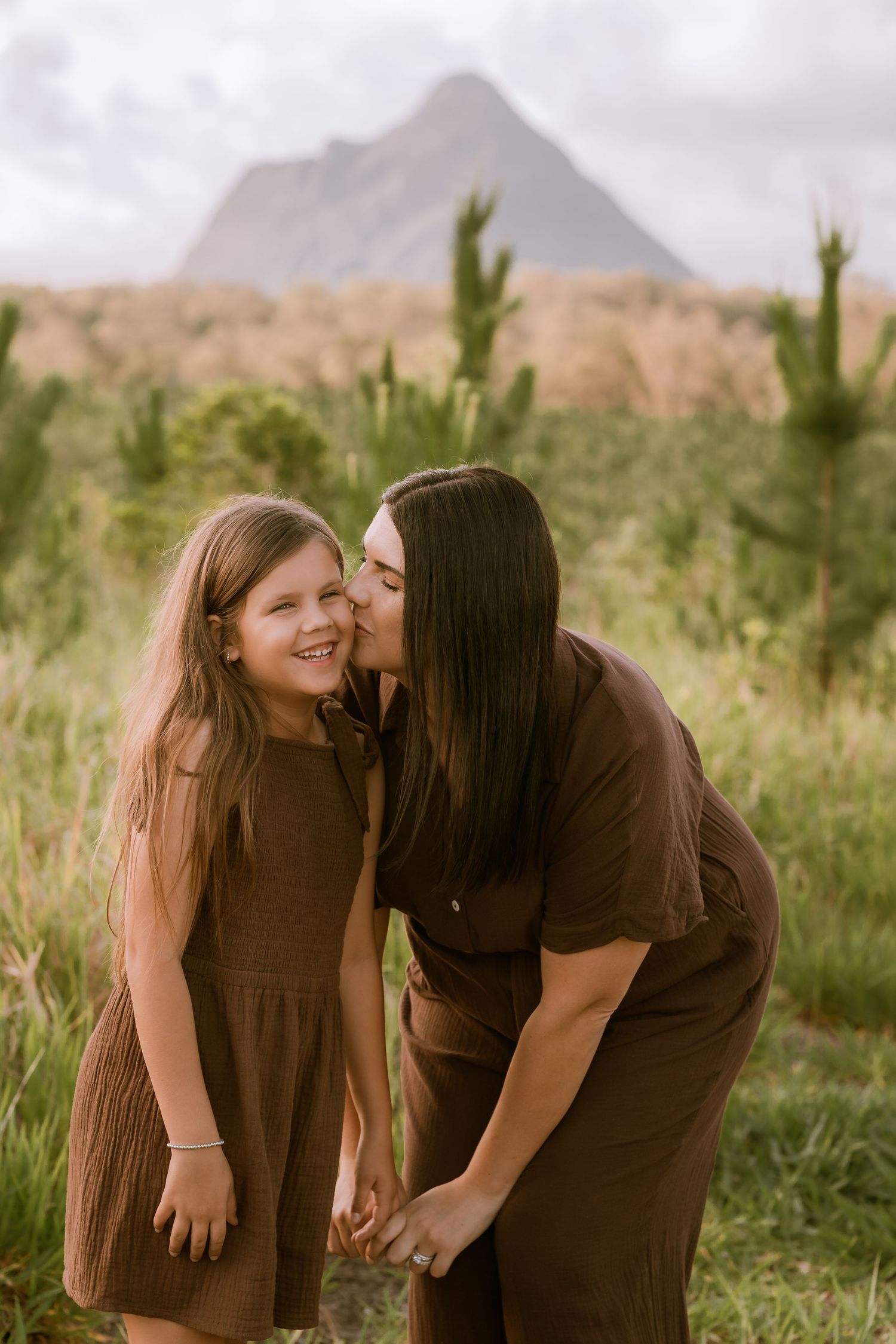 Brisbane family photographer — mother and teenage daughter laughing and sharing a tender moment in an open field with mountain backdrop — Jo