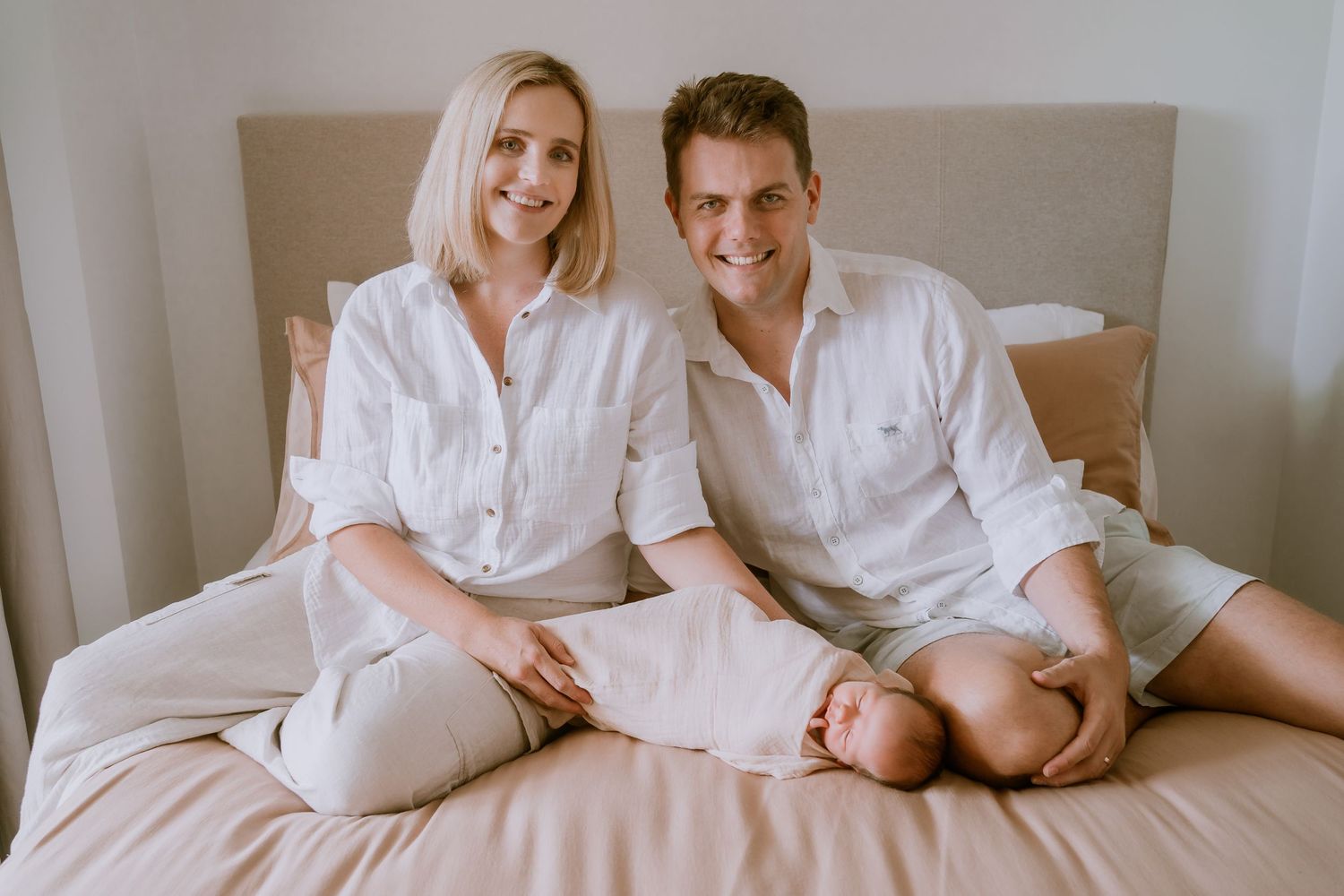 Brisbane newborn photographer — parents sitting together on a bed holding their newborn baby, warm and relaxed in-home lifestyle newborn ses