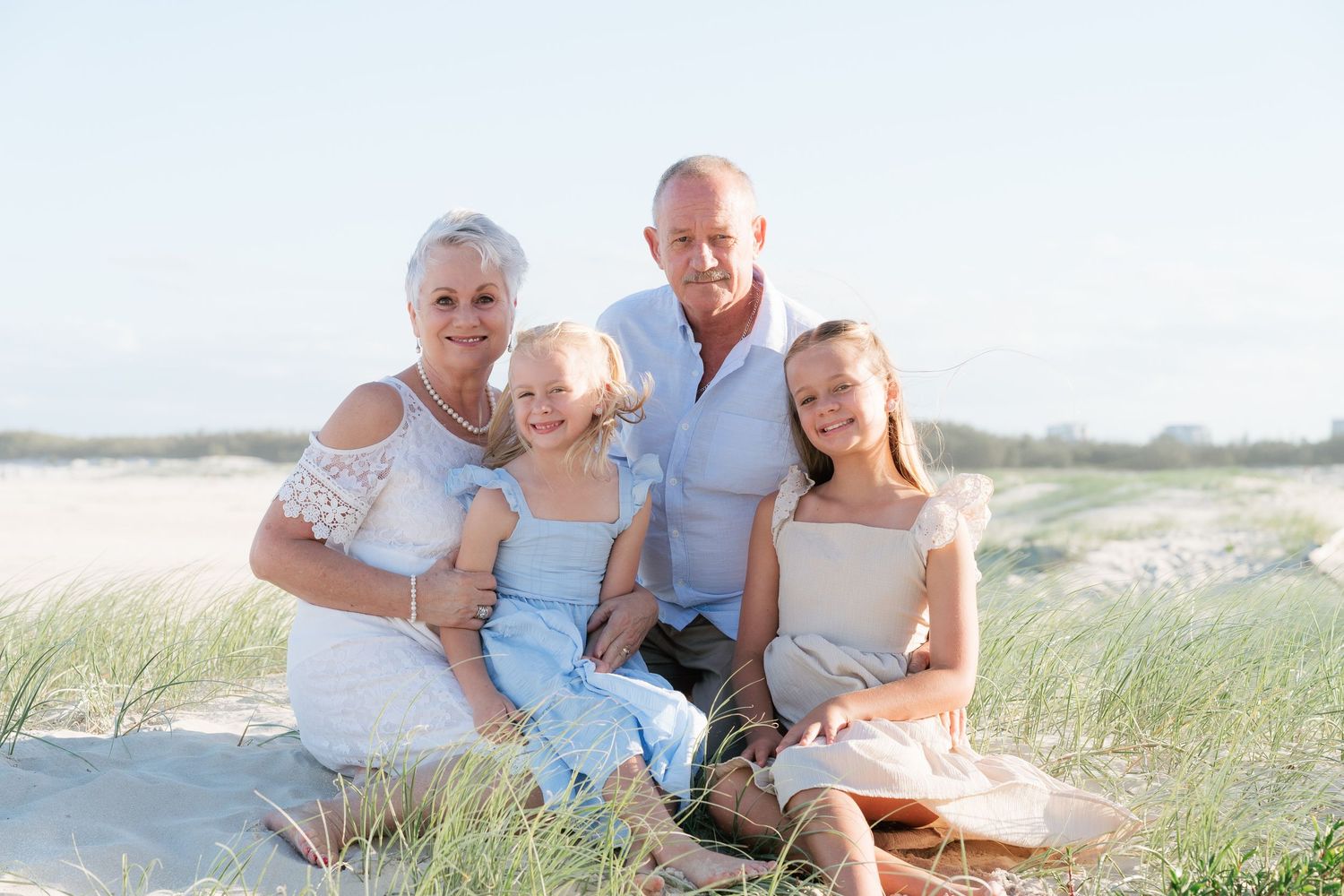 Brisbane family photographer — grandparents and two granddaughters sitting together on beach grass, relaxed multigenerational family portrai