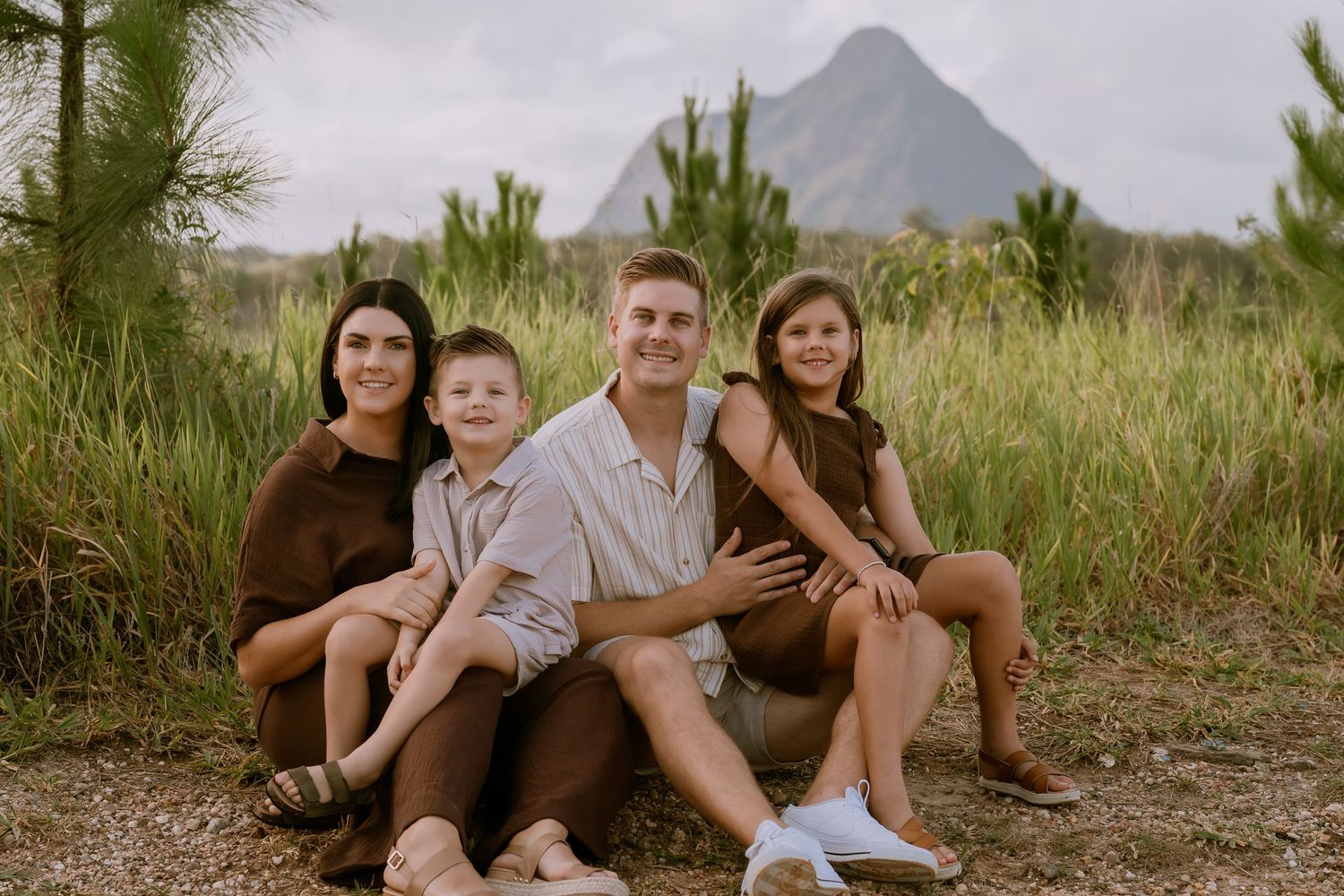 Brisbane family photographer — family of four sitting together in golden grass field with mountain backdrop, warm and relaxed lifestyle sess