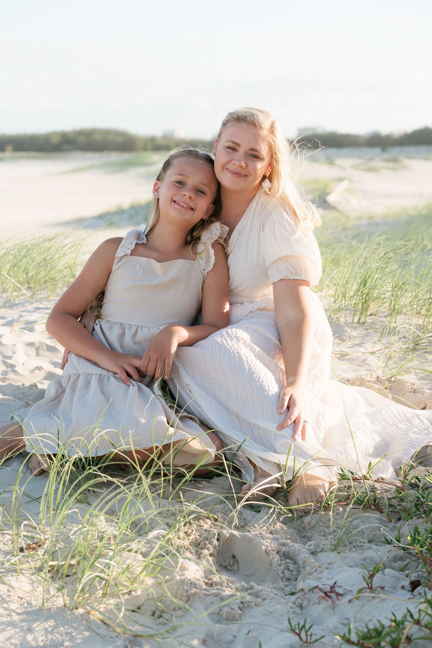 Brisbane family photographer — mother and daughter sitting together on sand dunes in matching white outfits, beautiful natural light beach s