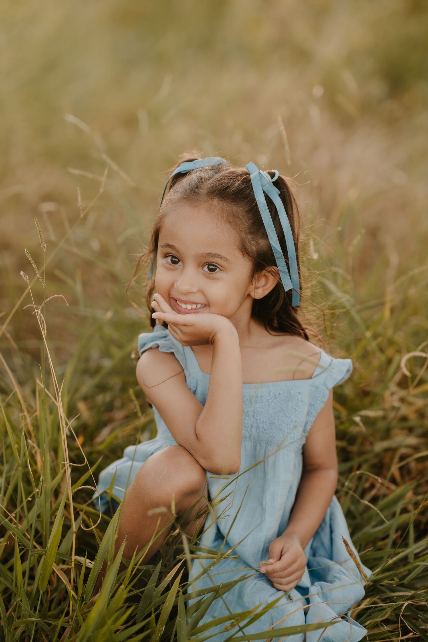 Brisbane family photographer — young girl in blue dress sitting and smiling in golden outdoor field, candid children's portrait by Joy Fille