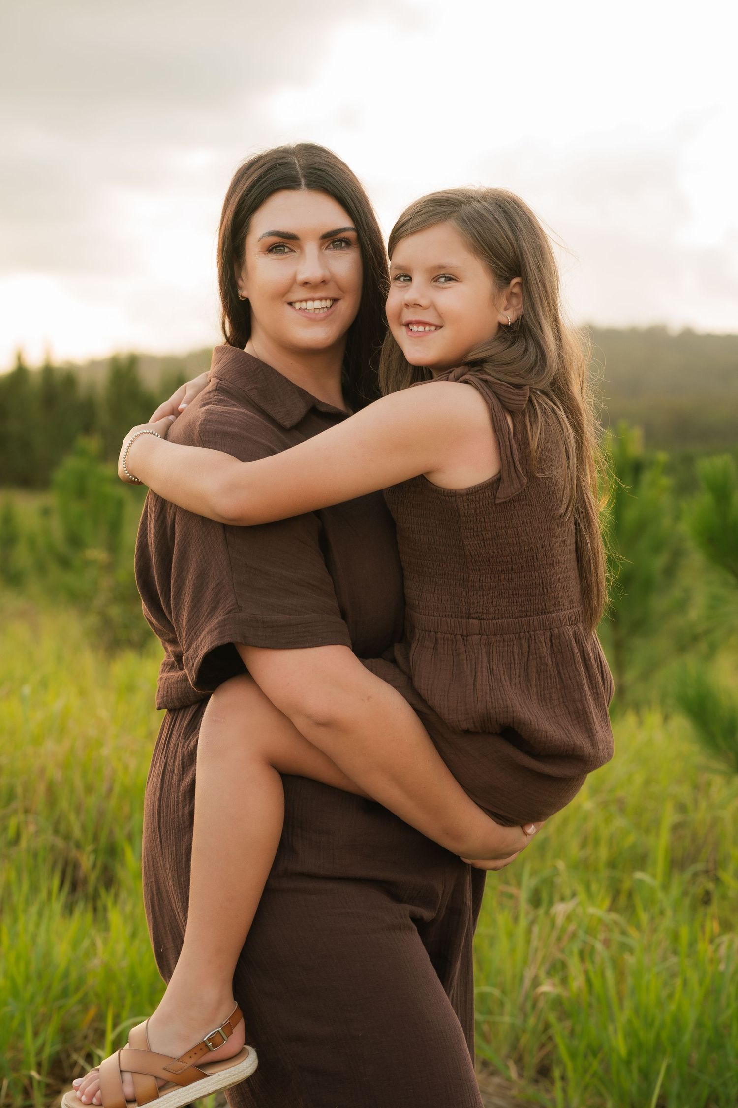 Brisbane family photographer — mother hugging daughter on her back laughing together in golden outdoor field, joyful family portrait by Joy