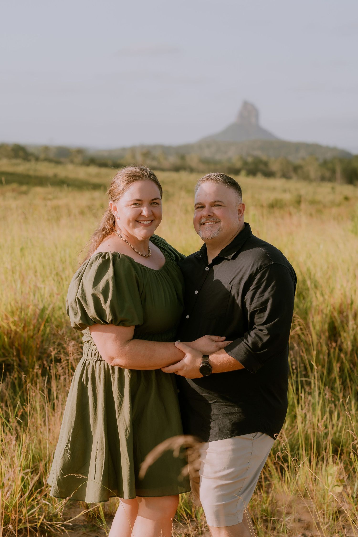 Brisbane family photographer — couple standing together in golden field with mountain backdrop, warm outdoor portrait by Joy Filled Photogra
