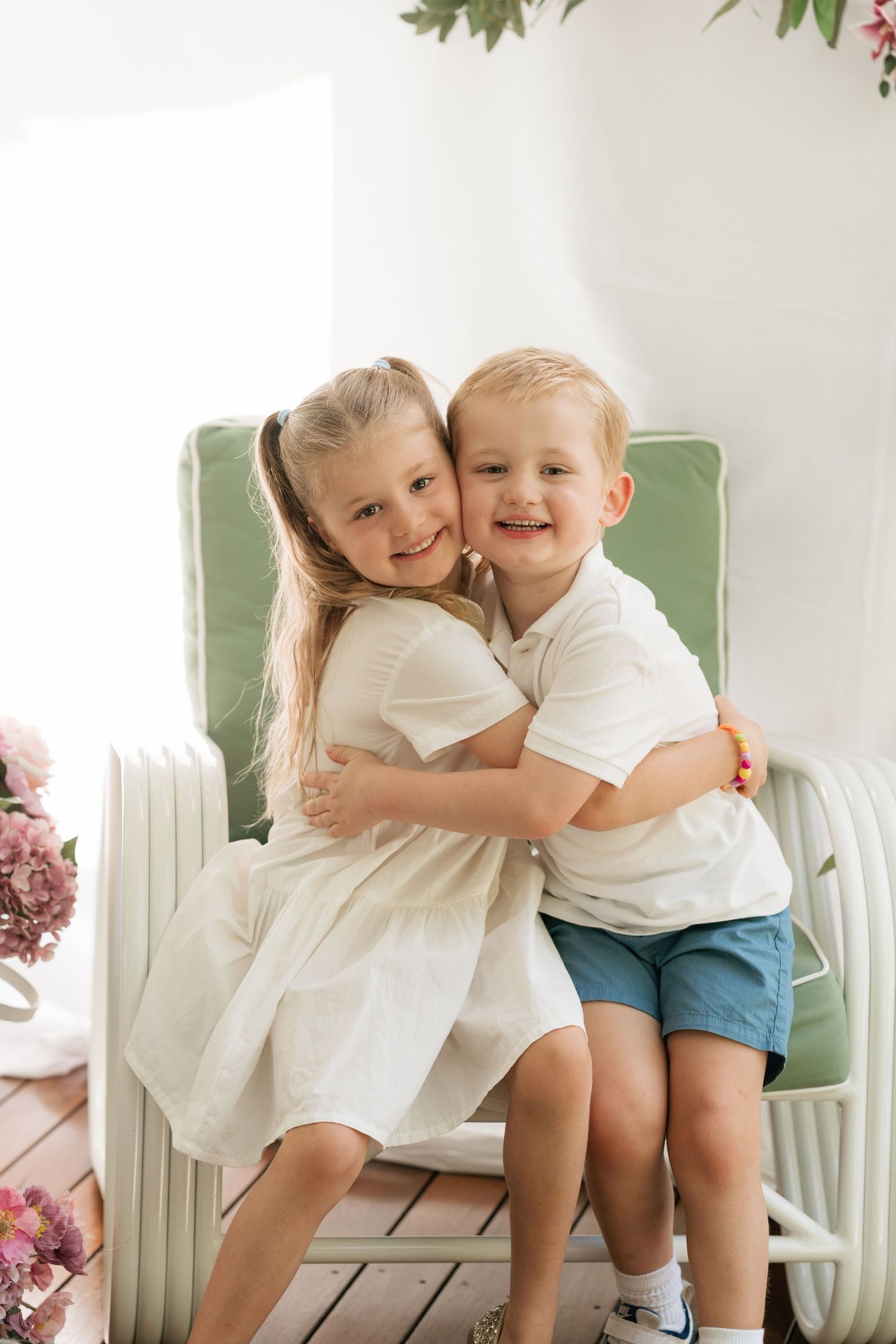 Brisbane family photographer — two young siblings hugging and smiling on a green chair indoors with floral decor, captured by Joy Filled Pho