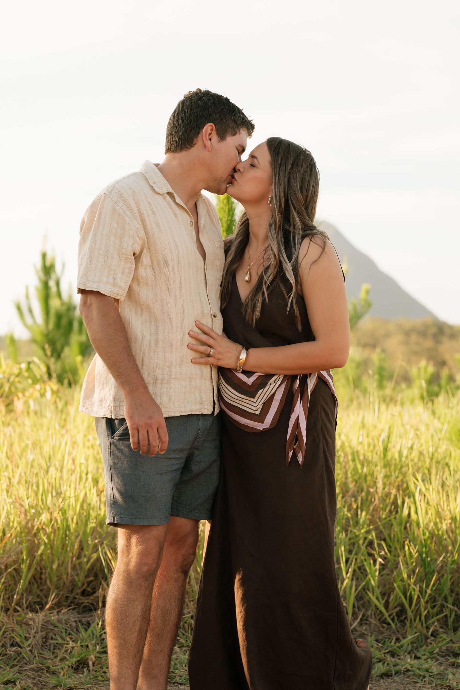 Brisbane couples photographer — couple sharing a romantic kiss at golden hour in lush outdoor grass, candid and intimate portrait by Joy Fil