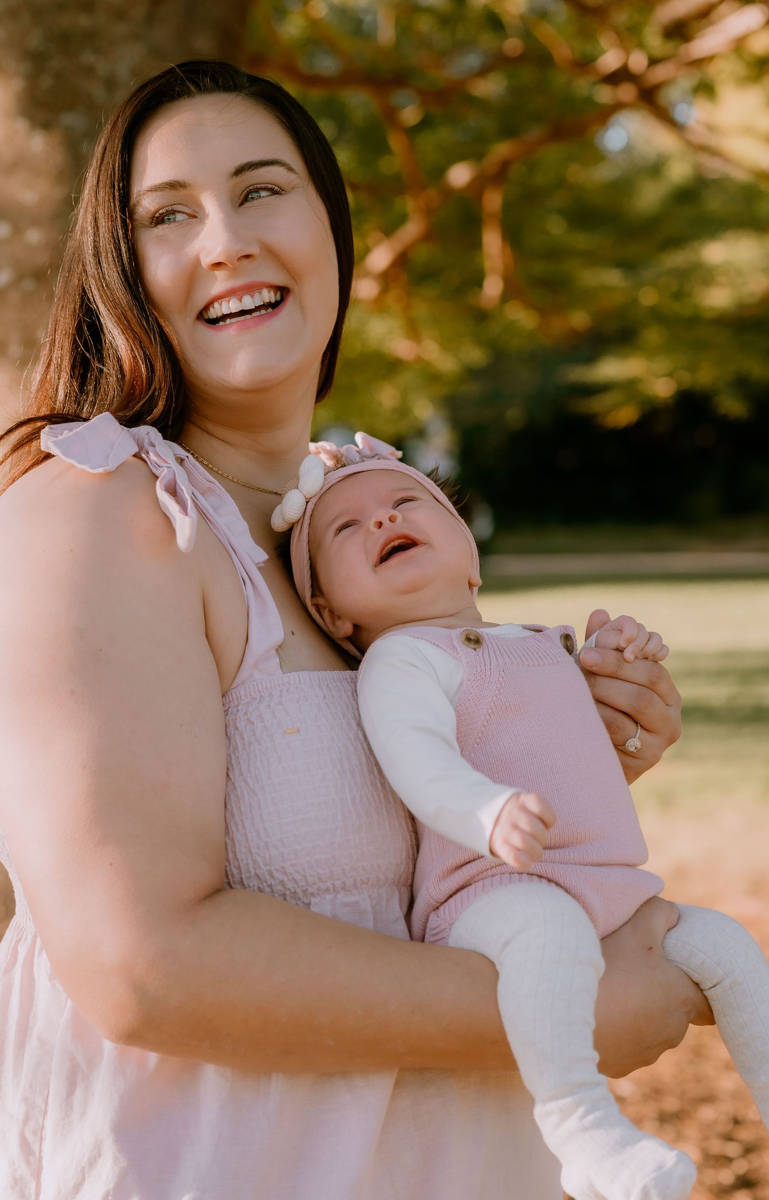 Brisbane family photographer — mother laughing joyfully while holding her baby in pink outdoors, genuine and heartwarming family moment by J