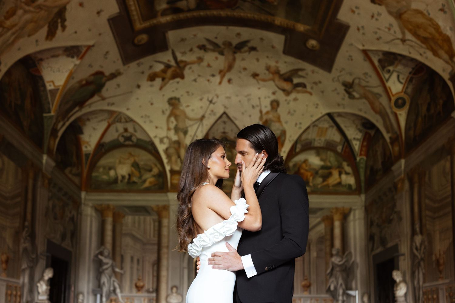 A romantic couple shares a kiss in an ornate baroque palace hall with painted frescoes and gilded decorations.