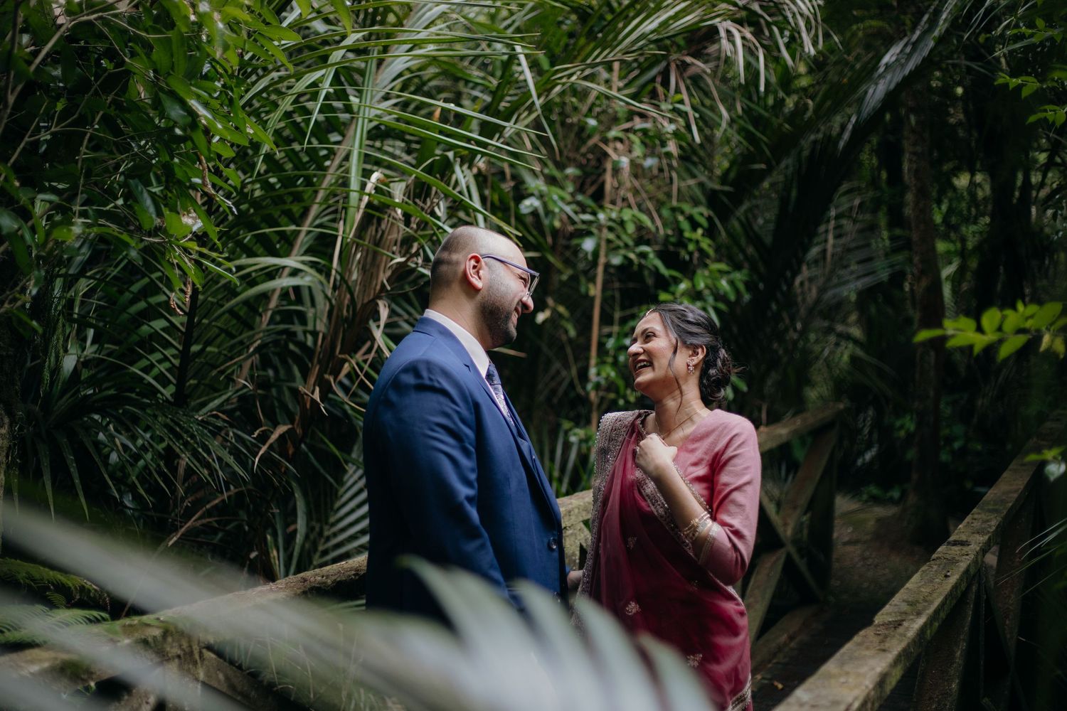 Two people share a joyful moment together while standing among lush tropical plants and palm fronds in a garden setting.
