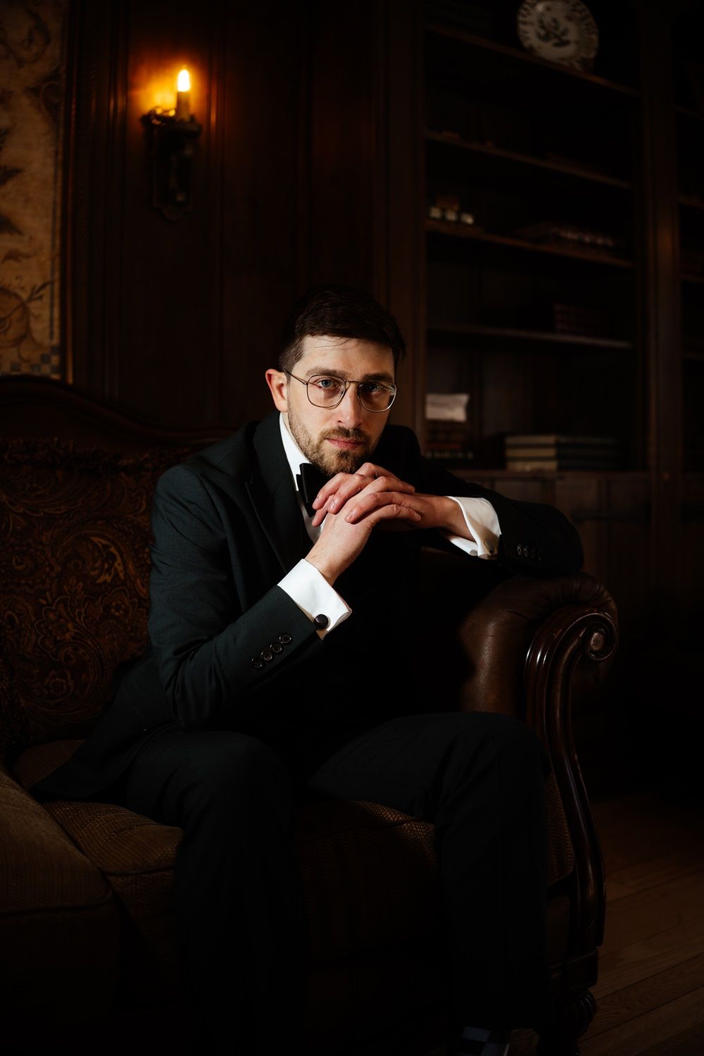 A moody portrait of a groom in a dark suit and glasses sitting thoughtfully in a vintage leather chair in Ontario.