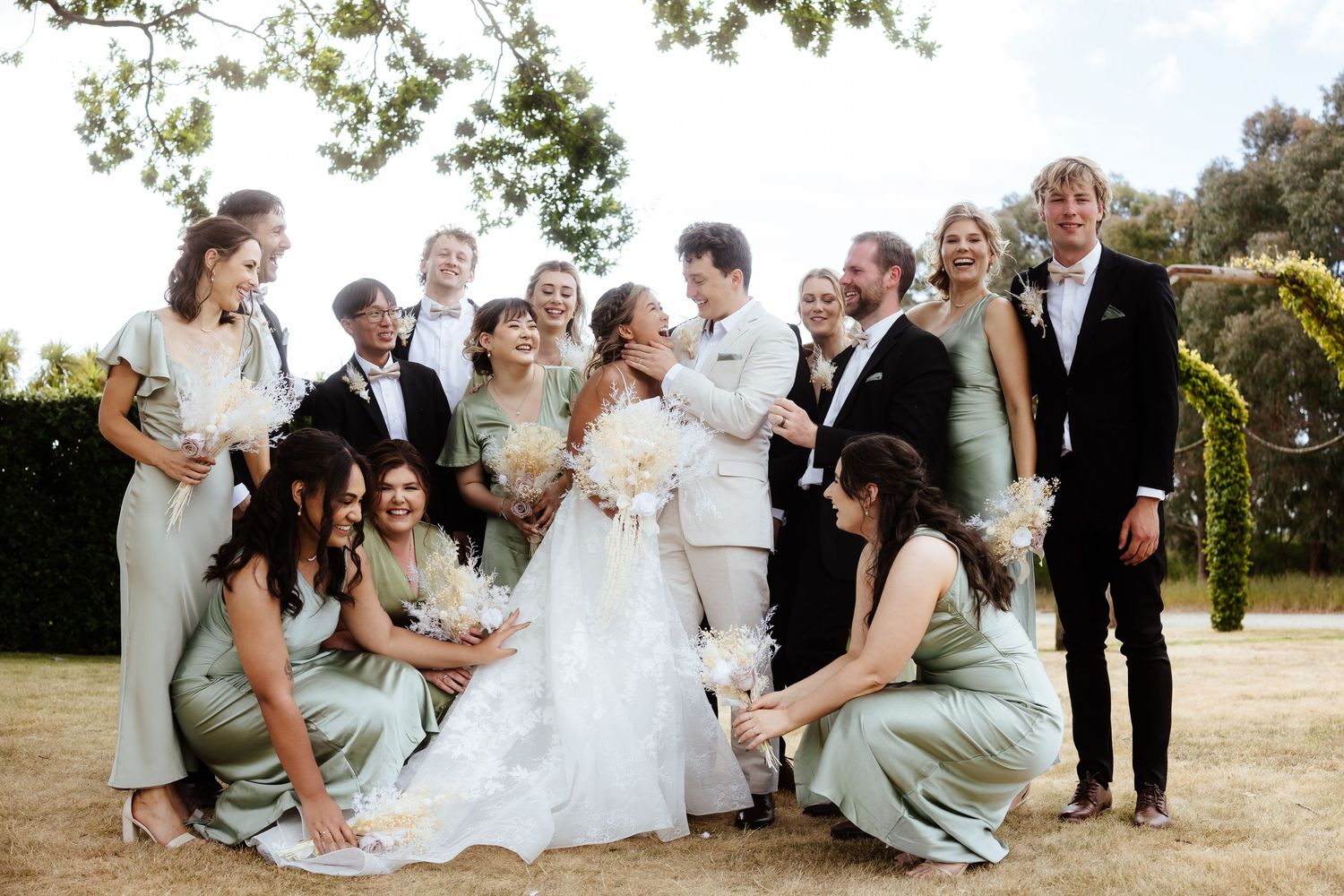 A joyful wedding party in light green bridesmaid dresses and formal attire poses together outdoors on a sunny day in Toronto.
