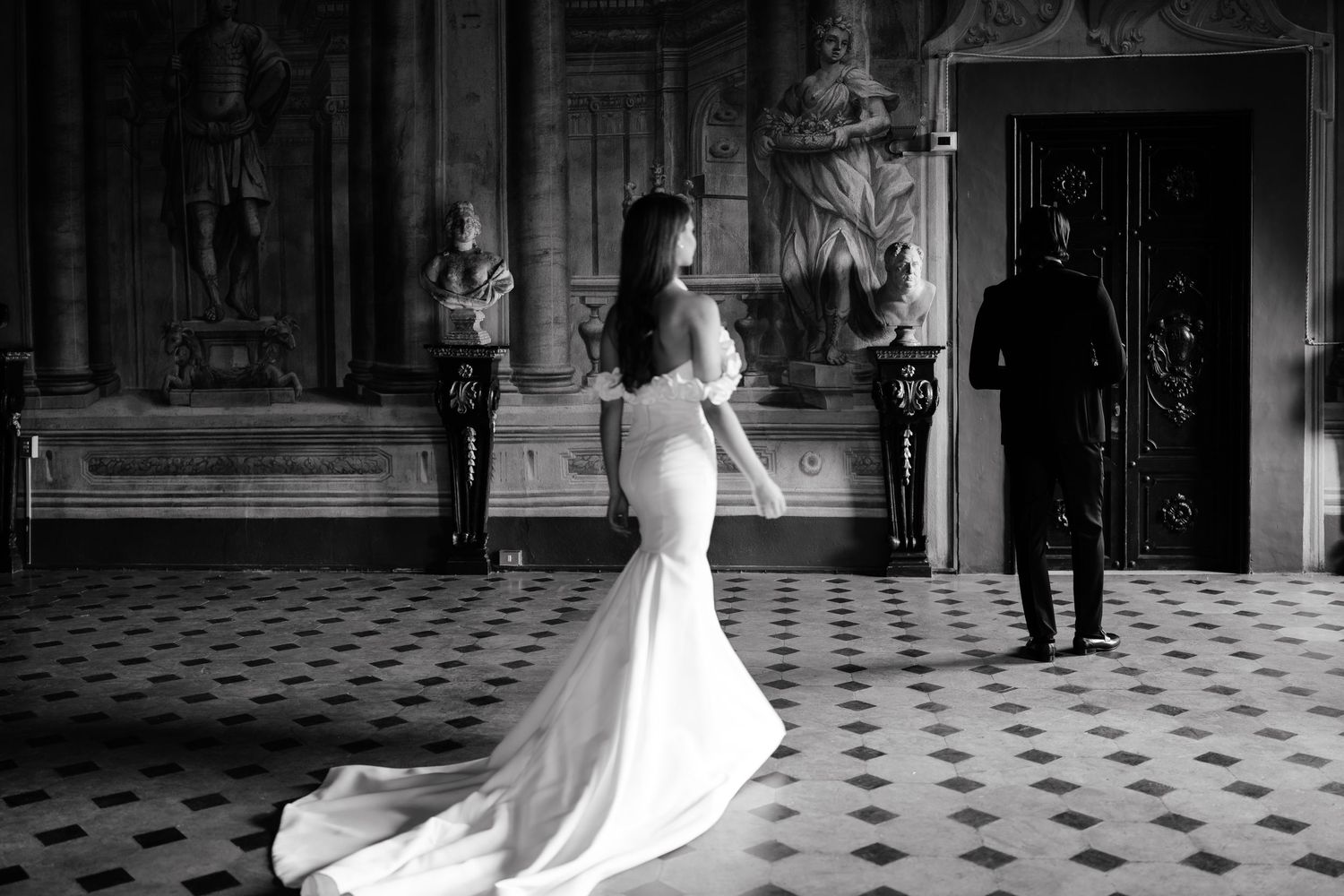 Black and white wedding photo showing a bride in a fitted mermaid gown with train standing in an elegant hallway.